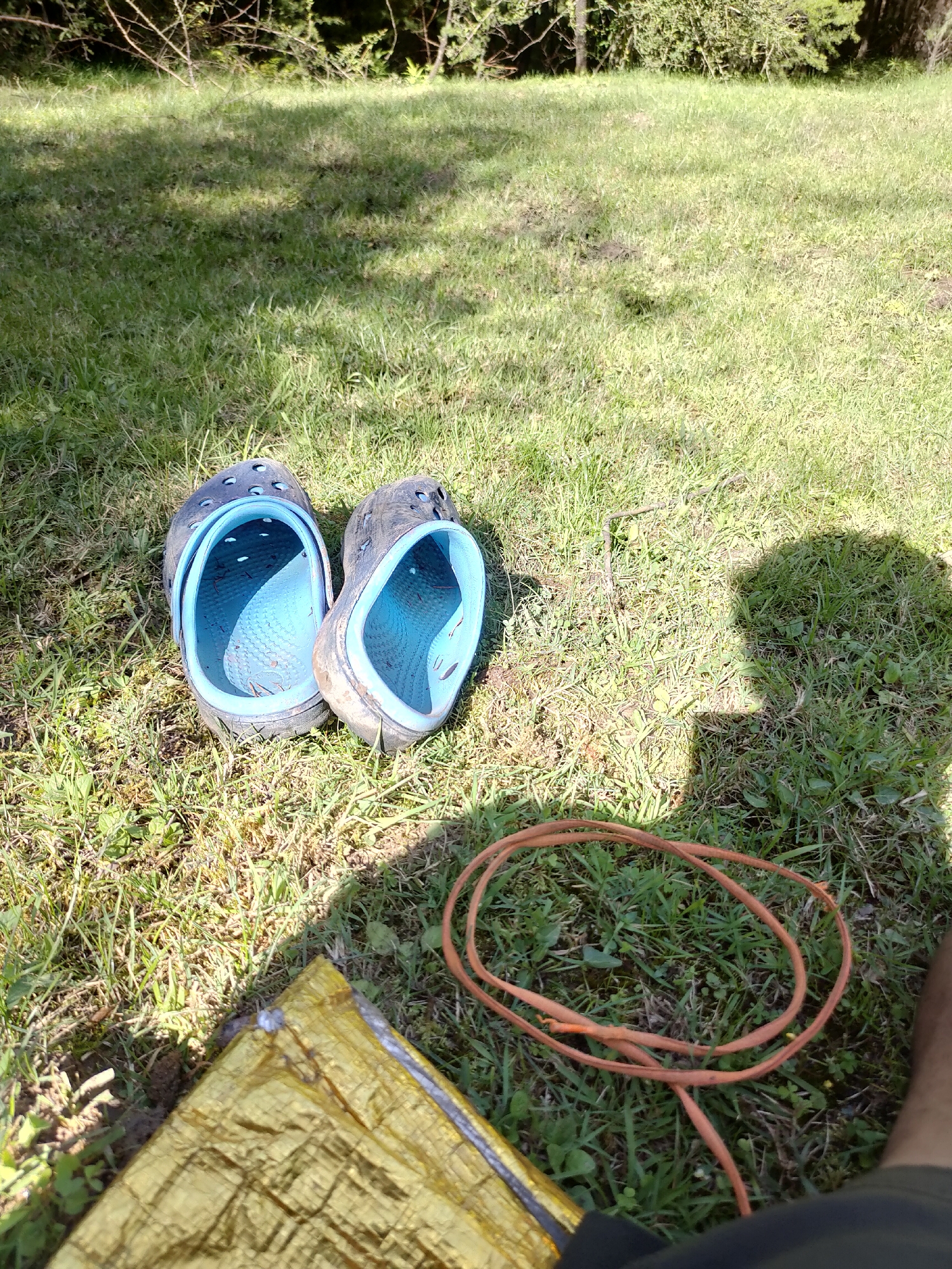 This is a medium-shot, eye-level, outdoor photograph taken in a grassy area with sparse trees in the background, possibly Manali, India. The image is primarily focused on the ground, featuring a pair of dark blue Crocs, one placed slightly in front of the other. Both shoes are facing upward and appear to be used. To the right of the Crocs, a coiled orange cord lies on the grass. In the lower left corner, a portion of a shiny, golden emergency blanket is visible. The ground is covered in green grass with some dry patches and fallen leaves. Sunlight casts shadows on the grass, suggesting it is daytime. A shadow of a person's head and upper body is visible on the right side of the image.