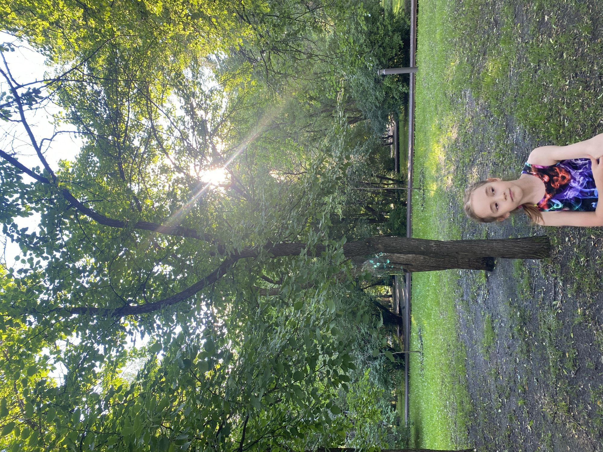 A young girl with blonde hair pulled into a ponytail, wearing a sleeveless multi-colored top, sits in a park. She is positioned on the right side of the frame, looking to her left with a neutral expression. The background features a lush green lawn interspersed with patches of dark soil and fallen leaves. Two trees stand prominently in the mid-ground, with sunlight filtering through their dense green foliage, creating lens flare effects. Beyond the trees, a pathway is visible, leading into a dense area of trees and greenery. The park appears to be in an urban setting, likely within the city of Yekaterinburg, Russia, given the context.