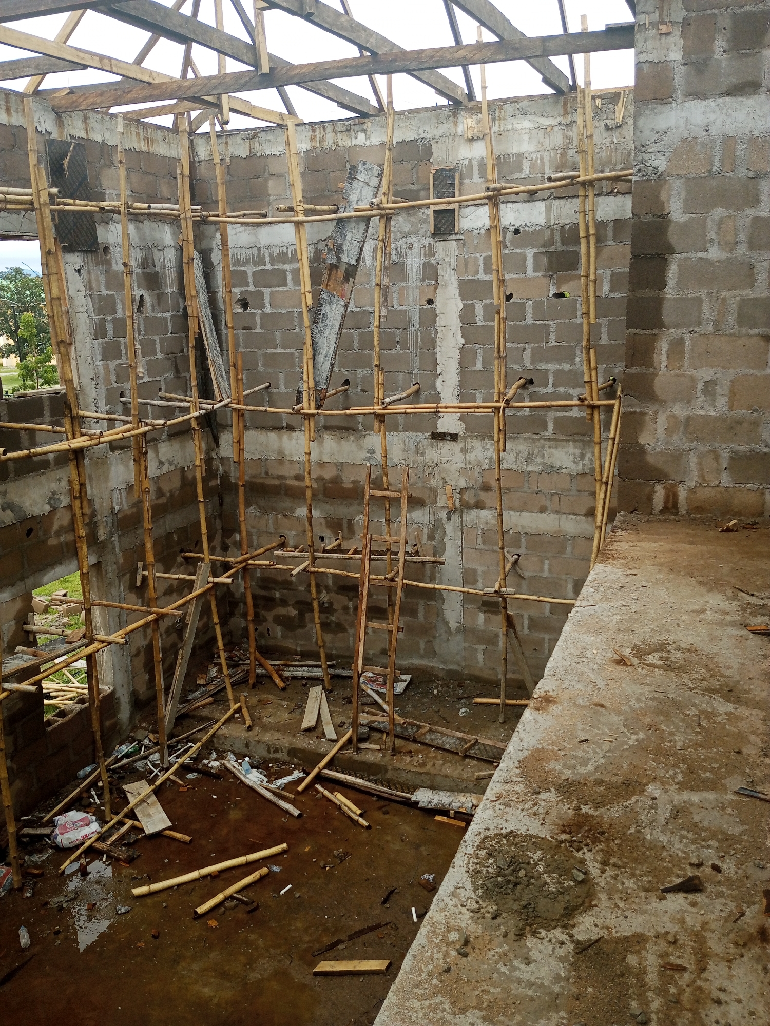 This media file captures an unfinished building construction site in Jalingo, Nigeria. The scene is characterized by raw, unplastered concrete block walls, forming the interior of a structure. Overhead, a skeletal wooden roof frame is visible, suggesting the early stages of construction.
The primary objects in the scene are the extensive bamboo scaffolding erected both inside and outside the building, supporting the structure and providing access for workers. Various construction materials are scattered on the muddy, wet ground, including wooden planks, bamboo poles, bags of what appears to be cement or building material, and some plastic debris. The floor of the building is uneven and covered in water and mud, indicating recent rainfall or ongoing work.
The lighting suggests it is daytime, possibly overcast due to the diffused light and lack of strong shadows. There are no people visible in the frame, focusing solely on the constructed elements and materials. The overall impression is one of ongoing construction, a temporary state of incompleteness. The presence of bamboo scaffolding is a notable detail, often used in construction in regions where it is readily available and cost-effective. No visible text is discernable in the image.