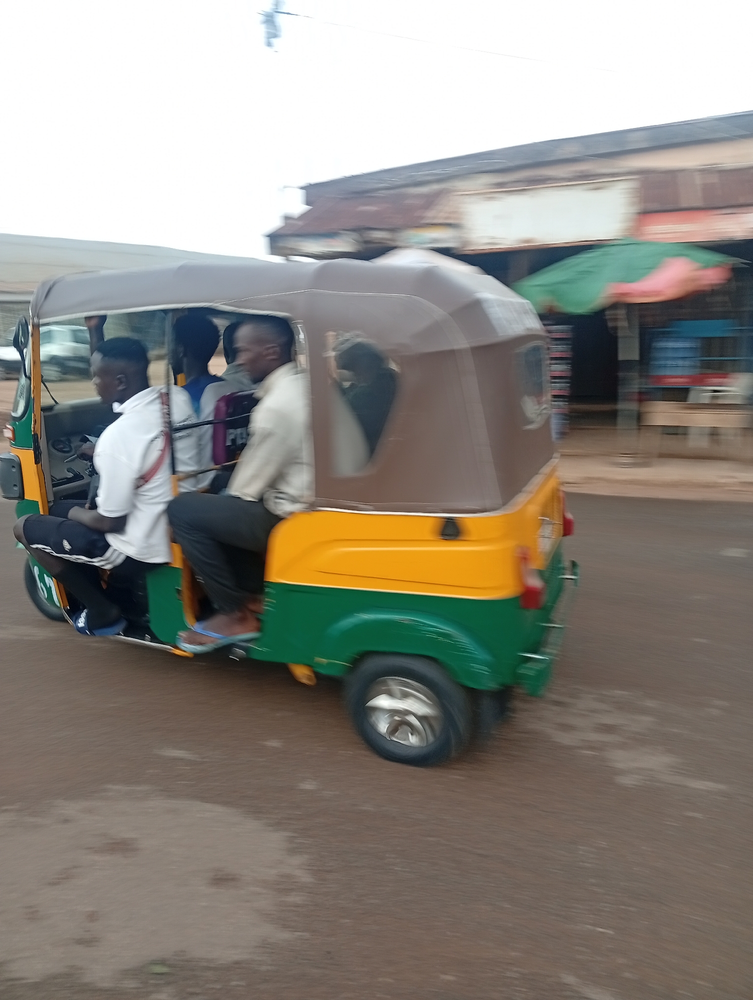 A vibrant green and yellow auto-rickshaw, commonly known as a Keke Napep, is captured in motion on a reddish-brown road in Jos, Nigeria. The vehicle features a brown canvas canopy and appears to be carrying multiple passengers.
In the front, a male driver, wearing a white short-sleeved shirt and dark shorts, is positioned at the wheel. Beside him, another male passenger, dressed in a light-colored long-sleeved shirt and dark trousers, occupies the front seat. He wears blue flip-flops. At least two additional passengers are visible in the rear compartment; their forms are partially obscured, but one person is seen wearing a greyish top.
The image exhibits significant motion blur in the background and on the road surface, indicating the auto-rickshaw is moving. On the right side of the frame, blurred commercial buildings with corrugated metal roofs and a faded signboard are visible, suggesting a busy street or market area. A green and pink market umbrella stands out against the background, near what appear to be water containers. The sky is bright and overcast, implying it is daytime. The scene depicts a typical mode of public transport in a bustling urban or semi-urban environment in Nigeria. No clearly legible text is discernible within the image due to distance and motion blur, although some indistinct letters (possibly "POL") can be vaguely made out on a rear passenger's shirt.