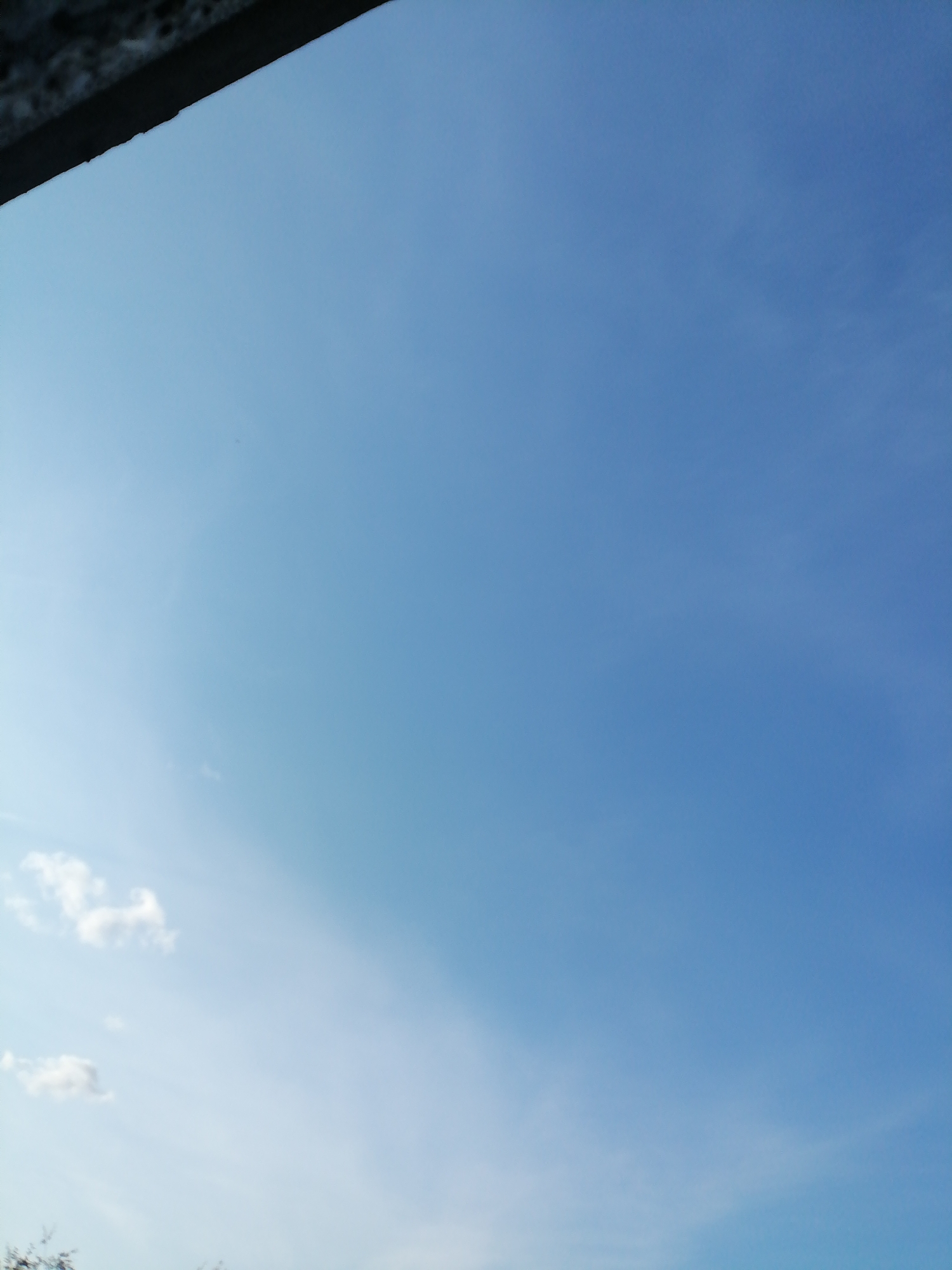 The image captures a clear blue sky with a few wispy clouds scattered across it. In the upper left corner, a dark, textured object, possibly a building overhang or a natural formation, partially obstructs the view of the sky. The lighting suggests it is daytime. The scene is devoid of any people or discernible man-made structures apart from the partial obstruction. The overall impression is of an outdoor environment with an expansive sky.