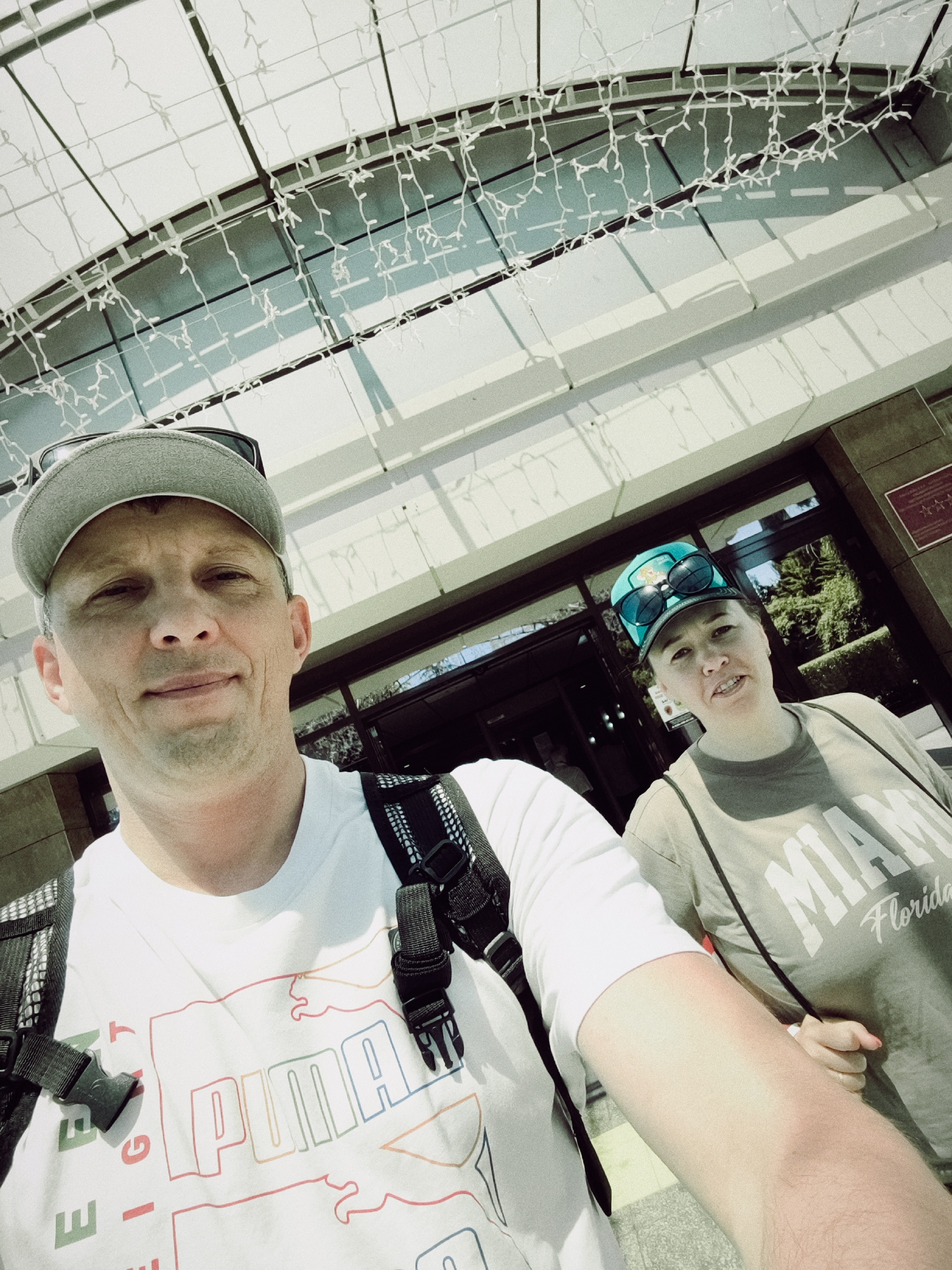 In Sochi, Russia, two individuals pose for a selfie in front of a modern building with a white, textured canopy ceiling adorned with string lights. The man in the foreground, wearing a white t-shirt with a colorful "PUMA" logo and a gray baseball cap, smiles at the camera. To his right, a woman wears a tan t-shirt with "MIAMI Florida" printed on it and a turquoise baseball cap with sunglasses resting on top. Behind them, the building features large glass windows and a dark entrance. The overall scene suggests a bright, sunny day, possibly at a hotel or resort entrance.