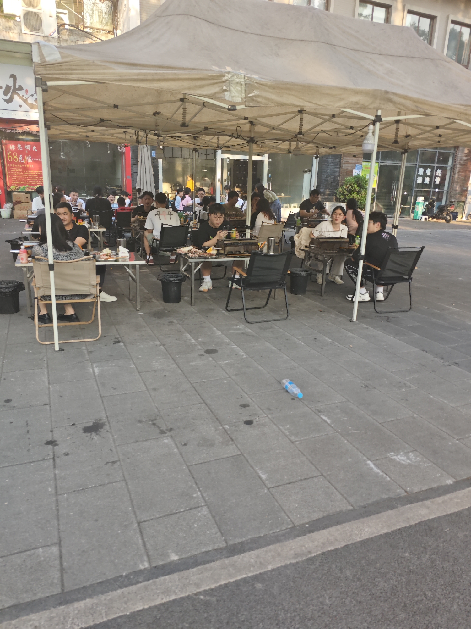 The image depicts an outdoor dining area in Chongqing High-tech Zone, China. A large, open-sided tent structure provides shade for multiple tables where people are seated and eating. People are interacting with each other, food, and grilling equipment. Tables are arranged with chairs around them, with garbage cans placed nearby. A discarded plastic bottle lies on the paved ground in the foreground. The setting appears to be a commercial area, with buildings and some vegetation visible in the background.