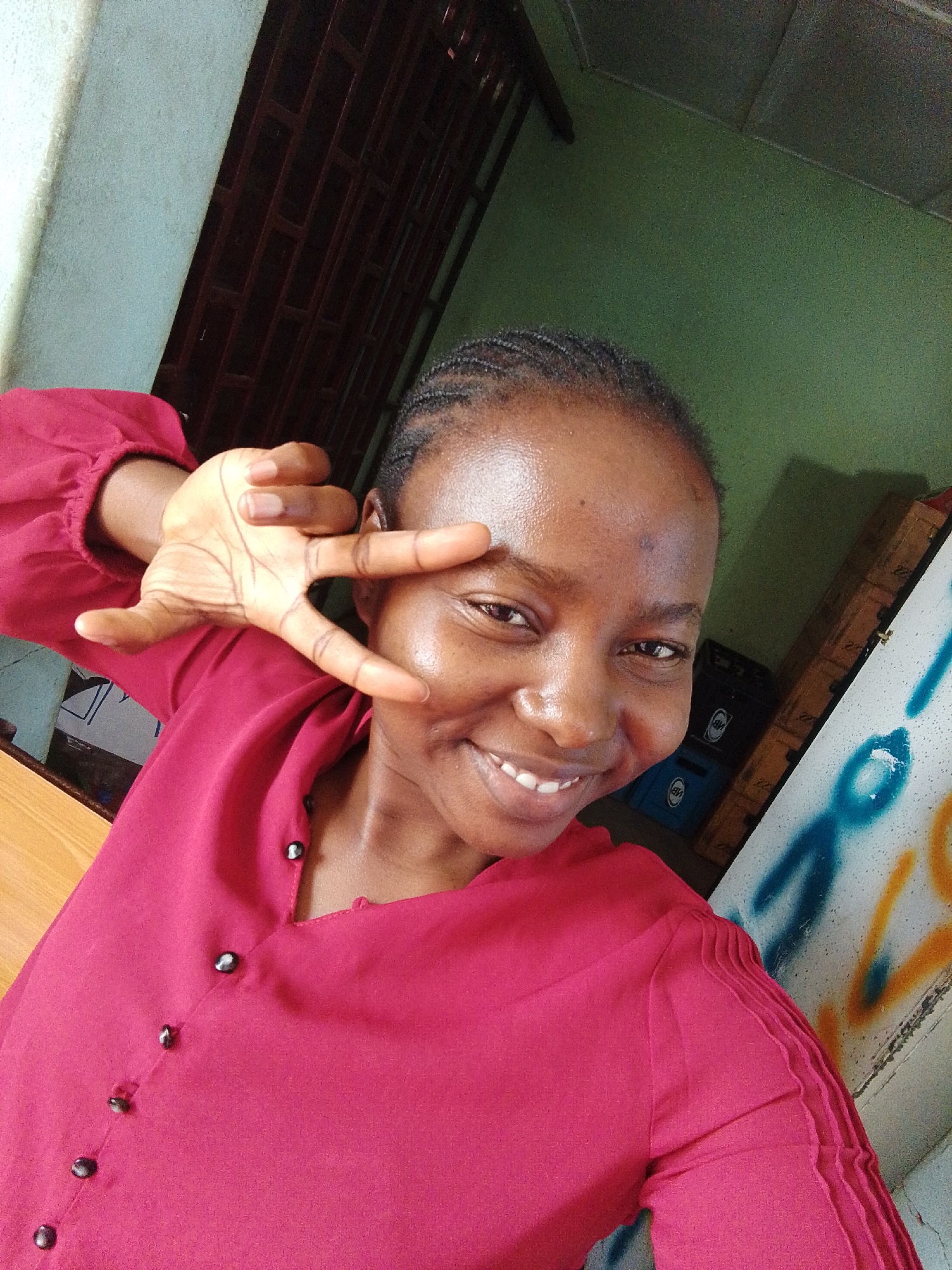 The image shows a woman in Unknown, Unknown taking a selfie indoors. She is smiling and holding up a peace sign with her left hand. She is wearing a red, long-sleeved button-down shirt. Her hair is braided. Behind her, there is a wall painted green, a brown door with a grid pattern, and a white surface with blue and yellow graffiti. There is also a stack of blue crates visible in the background.