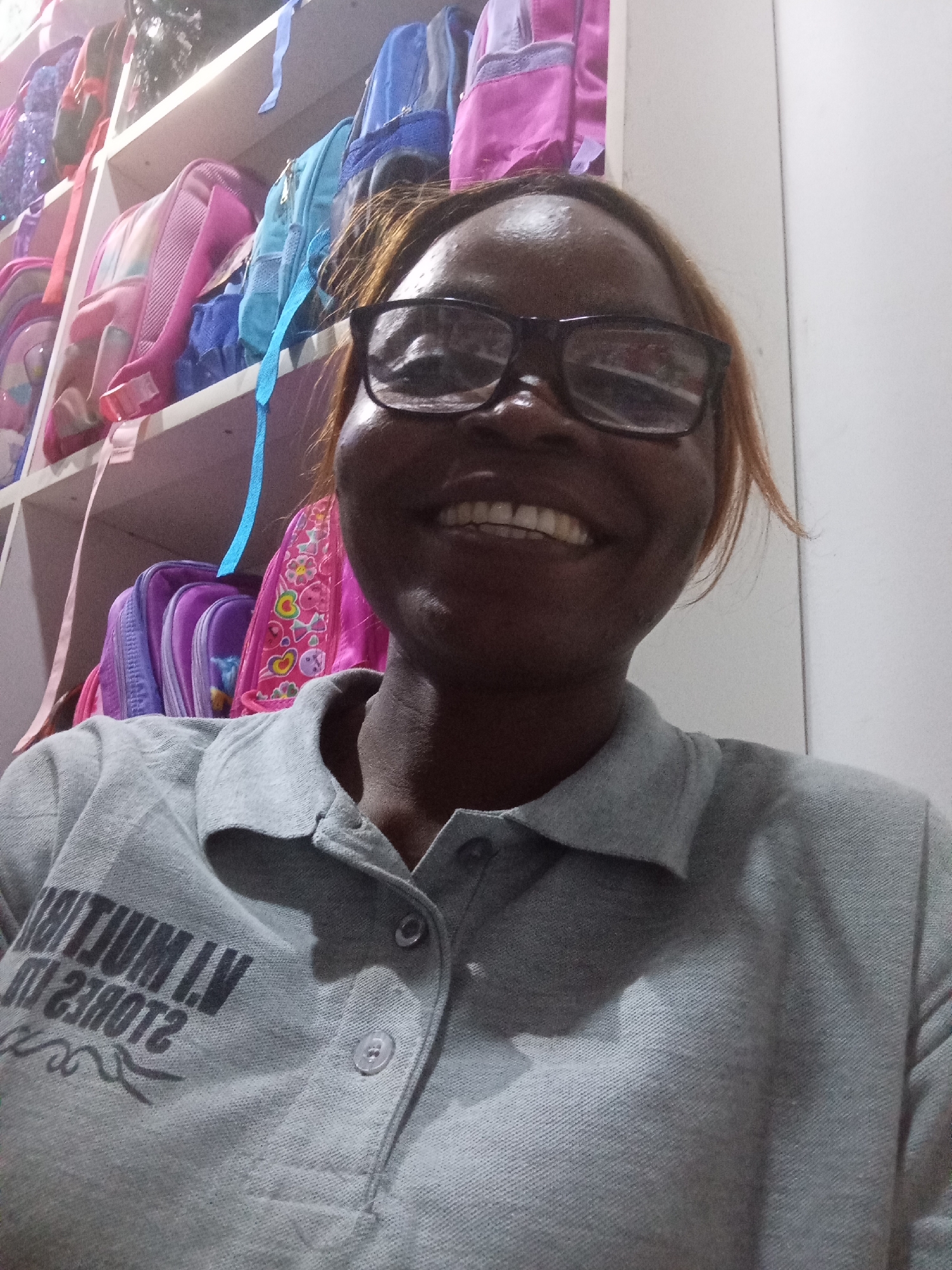 A medium shot captures a woman with dark skin, brown hair, and glasses smiling at the camera. She is wearing a gray polo shirt with a logo that includes the text "VNTUM LV" and "210812". Behind her, on shelves, are numerous colorful backpacks. The background is out of focus, but suggests an indoor setting, potentially a store or a room filled with backpacks. The image is lit from the front, casting subtle shadows. The location is the city of Howayi, Nigeria.