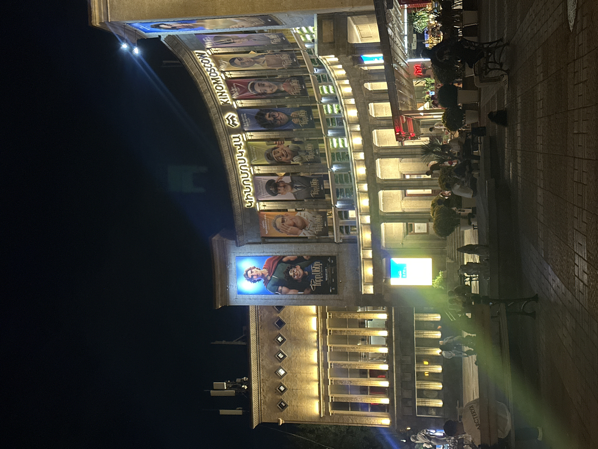The image shows a street scene at night in Yerevan, Armenia, likely in the city center due to the presence of a cinema. The main subject is the illuminated facade of a cinema building, identified as "KINOMOSCOW" by banners and signage. Several large movie posters featuring animated characters and possibly human actors are displayed prominently on the curved facade. The building itself is multi-story, with extensive lighting highlighting its architectural features, including columns and arched windows. In the foreground, a public square with brick paving is visible. Several people are present in the square, some sitting on benches or at tables outside a cafe or restaurant area attached to the cinema. The overall atmosphere is lively and urban, typical of a city at night.