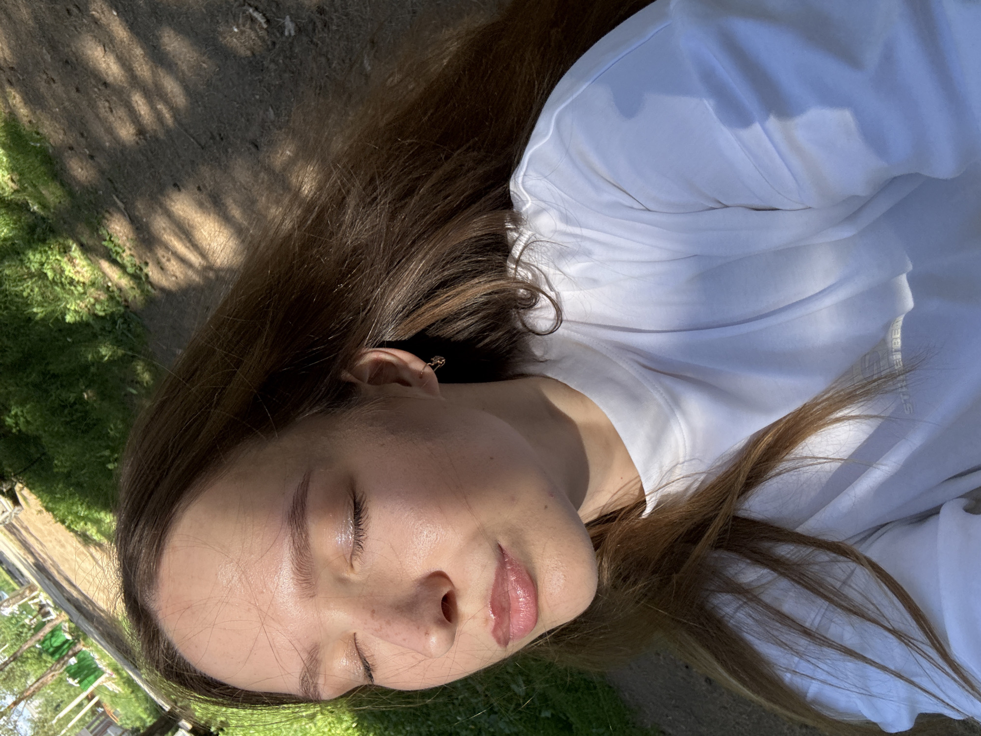 A young woman with long, brown hair is captured in a close-up selfie outdoors. She is wearing a white t-shirt and has her eyes closed. Sunlight is illuminating her face, highlighting her features and the texture of her skin. She is wearing a small earring in her right ear. The background shows a path with dappled sunlight and shadows, suggesting a park or a tree-lined area. The presence of green grass and trees indicates a natural environment within the city of Chaikovsky, Russia.