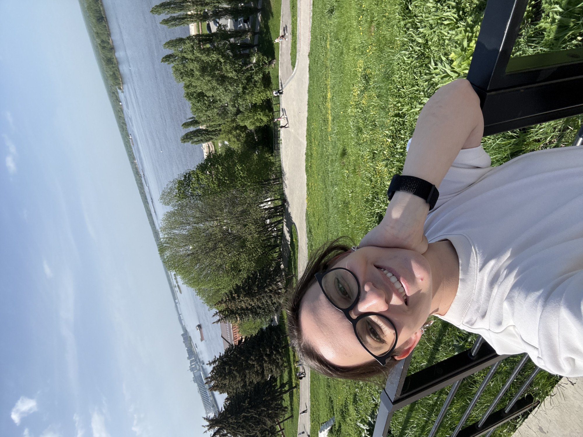 A selfie of a woman with dark hair and glasses, wearing a white shirt and a black watch, is taken from an elevated point. She is smiling and leaning against a black metal railing. In the background is a body of water, presumably the Volga River, with trees and greenery lining the banks. A paved pathway is visible running alongside the grassy area. The setting is likely a park or overlook in Nizhny Novgorod, Russia, given the distinctive river and cityscape features, with the possible location being the Verkhne-Volzhskaya Embankment.
