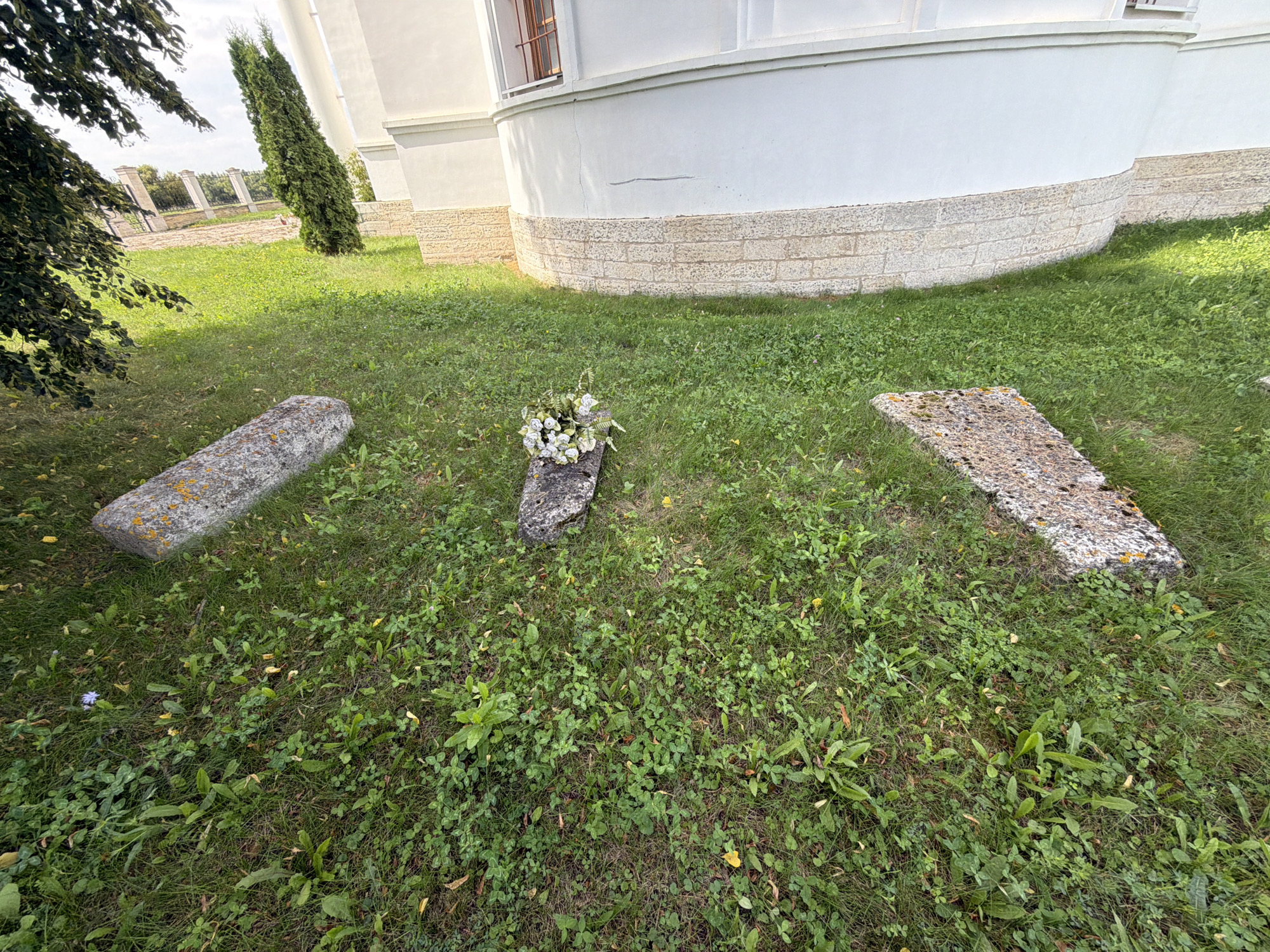 The image shows a grassy area in front of a large white building. Three rectangular stone slabs lie on the grass. The slab in the center has a bouquet of white flowers placed on it. The building appears to be a church or a similar historical structure, with a rounded facade and a base constructed of light-colored stone blocks. A tall, slender green tree stands near the building. The area is well-lit by natural daylight, suggesting it is daytime. The presence of the stone slabs and the flowers indicates a possible memorial or grave site. The scene is located in the village of Skornyakovo, Russia.