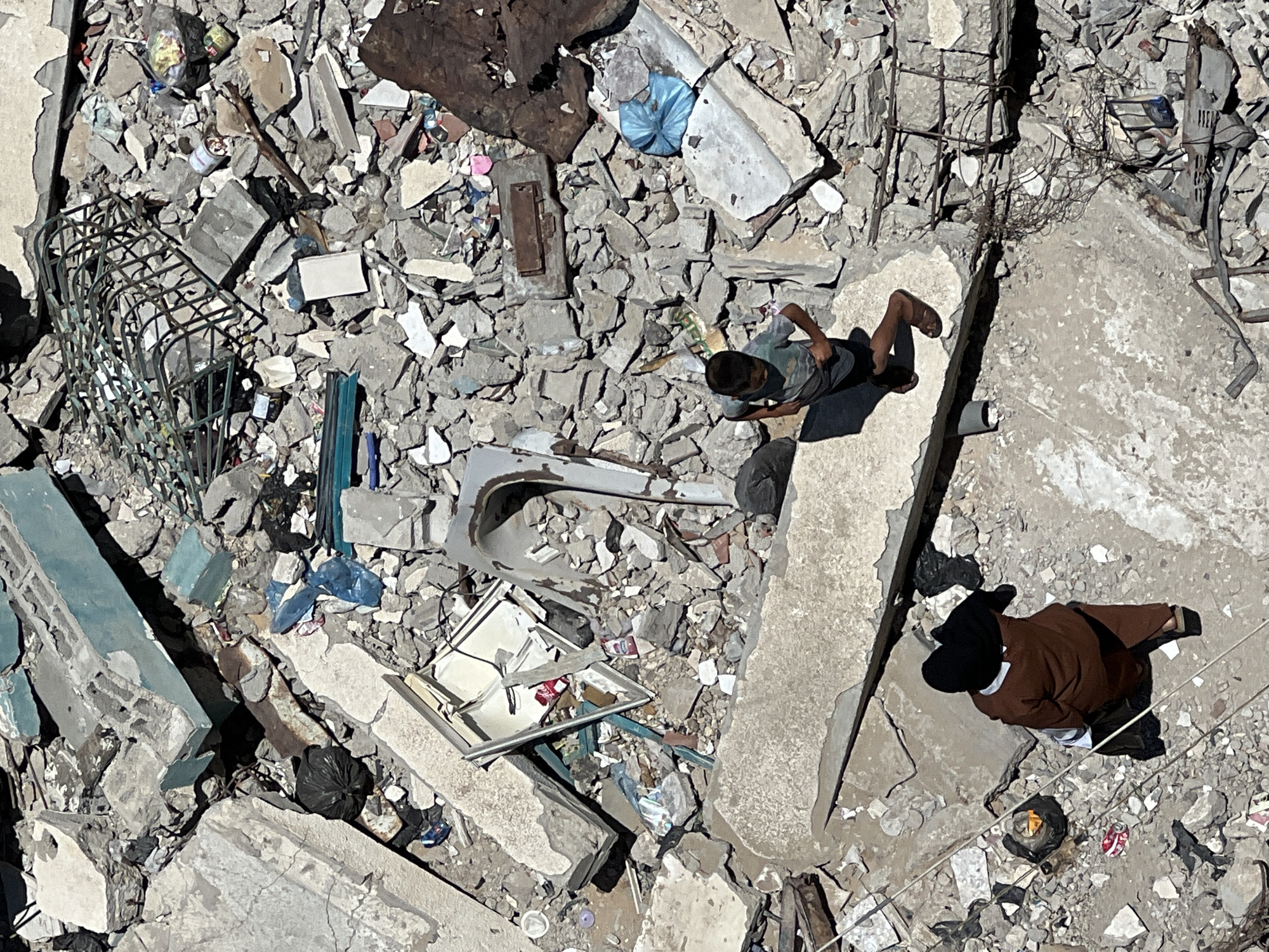 An aerial, high-angle medium shot shows a boy and an adult in a debris-filled urban environment in the city of Gaza, Palestinian Territories. The ground is covered in concrete rubble, twisted metal, and discarded items, indicating a scene of destruction. A young boy, wearing a grey t-shirt and dark shorts, is walking on a large concrete slab, his body angled to his left. To his left and further down the slab, a person wearing a brown abaya and a black hijab is walking away from the camera. The background is a chaotic jumble of broken building materials, including large concrete blocks, rebar, and what appears to be parts of furniture and appliances. Scattered amongst the debris are plastic bags, bottles, and other refuse. The scene is illuminated by bright sunlight, casting harsh shadows.