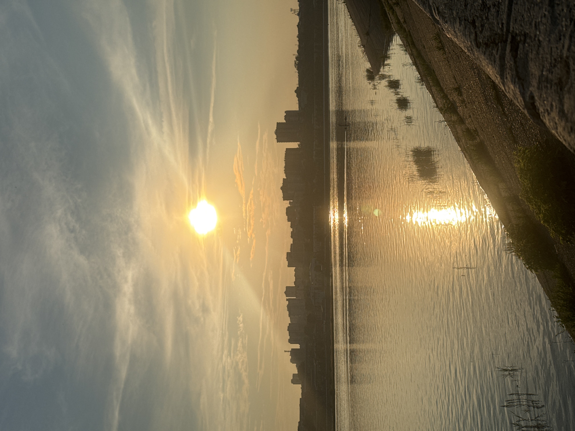 The image depicts a sunset over the Angara River in Irkutsk, Russia. The sun, a bright disc, is positioned in the upper center of the frame, casting a warm, golden light across the sky and water. Wispy clouds streak across the sky, illuminated by the setting sun.
In the middle ground, a city skyline stretches across the horizon, silhouetted against the bright sky. The buildings are a mix of modern high-rises and older structures, suggesting an urban landscape. The Angara River occupies the foreground and middle ground, its surface rippling with the reflection of the sun and the city lights. A man is visible on a stand-up paddleboard in the river.
The right bank of the river is visible, featuring a sloping embankment covered with vegetation and some concrete structures. The overall impression is one of a tranquil evening scene in an urban environment.