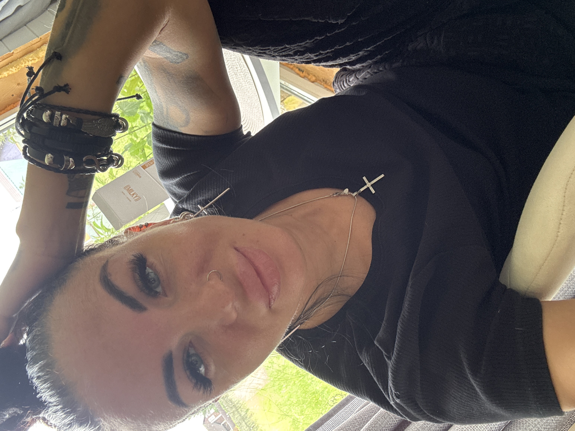 A close-up selfie shows a woman with dark hair pulled up in a bun, light blue eyes, and dramatic makeup. She is wearing a black t-shirt, a silver necklace with a cross pendant, and multiple bracelets on her left wrist. A nose ring is visible. Tattoos are present on her arms. She is indoors, near a window, with greenery visible outside. A small white box with text is also in the background. The setting appears to be a residential area.