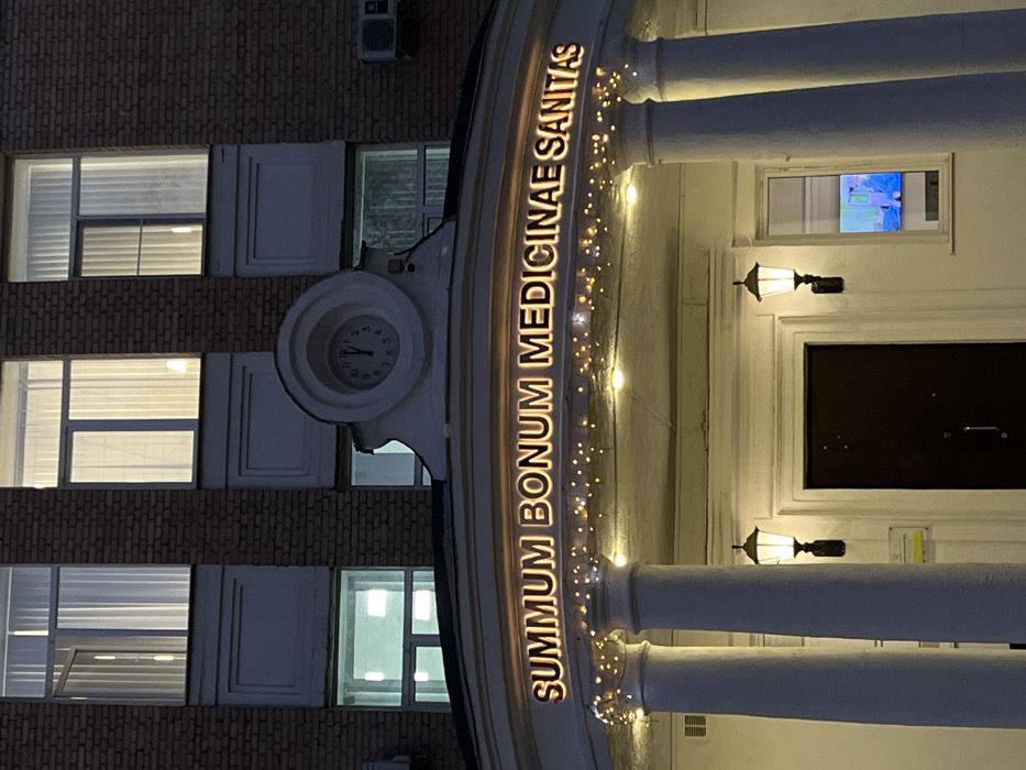 The image displays the illuminated facade of the main building of Sechenov University (First Moscow State Medical University named after I.M. Sechenov) in Moscow, Russia, at dusk or night. The brick upper stories feature multiple windows, some with interior lights visible and white curtains, one window partially ajar. Below, a prominent white classical-style portico is supported by two large columns. Along the arch of this portico, the Latin inscription "SUMMUM BONUM MEDICINAE SANITAS" (The highest good of medicine is health) is visible in illuminated golden letters, with decorative string lights draped beneath. Above the portico, a large clock set within a white architectural element indicates approximately 11:58. Two traditional-style lanterns illuminate the area around a dark, recessed main entrance door. To the right of the entrance, a ground-floor window reveals a brightly lit interior where a figure in blue medical scrubs is visible. Air conditioning units are mounted on the brick sections of the building. No other individuals are present in the exterior scene.