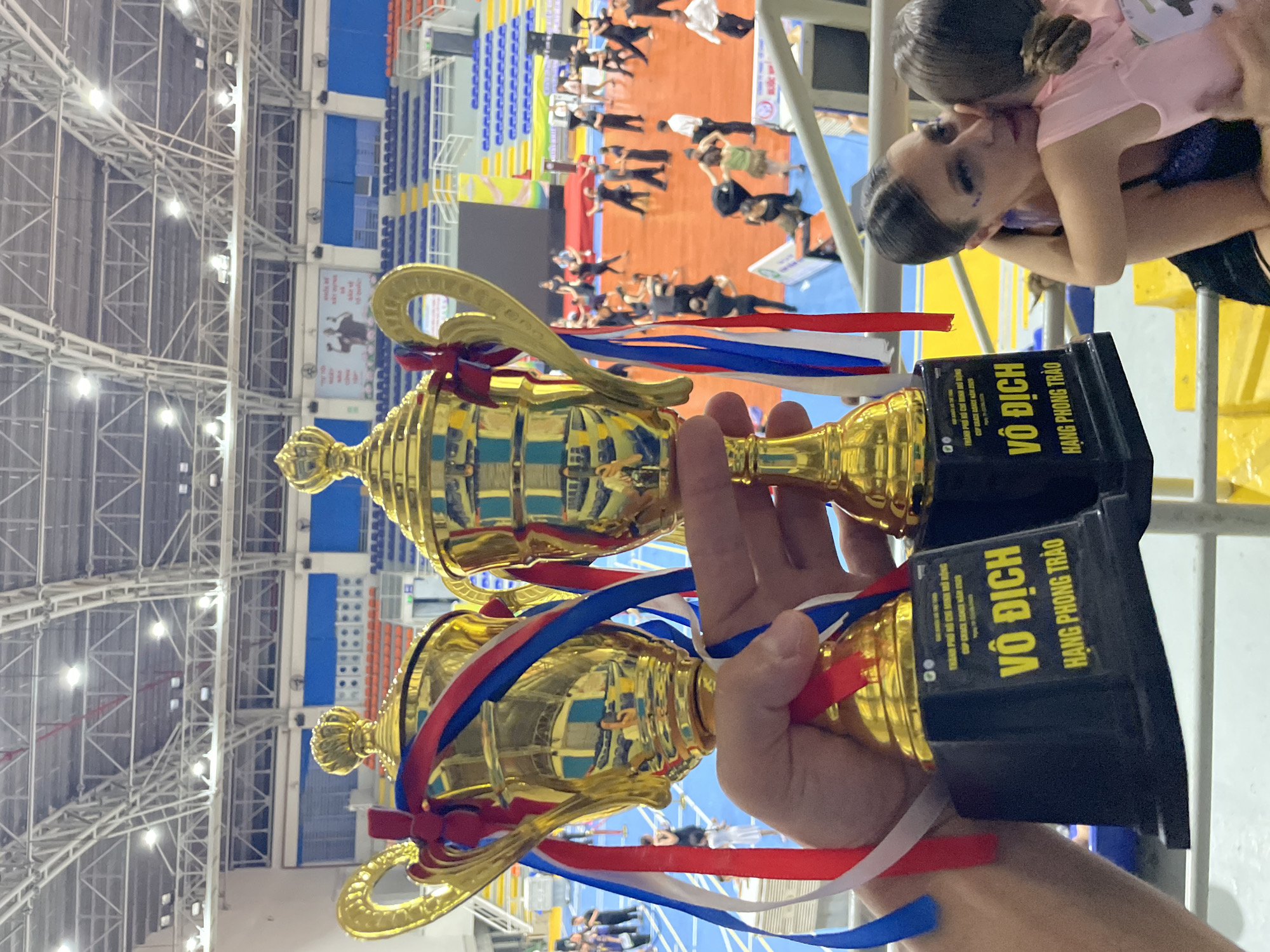 In a sporting arena, two gold trophies with red, white, and blue ribbons are held in the foreground by a person's hands. The trophies have black bases with yellow text that reads "VÔ ĐỊCH HANG PHONG TRÀO." In the background, a group of people in black and white attire are dancing on a red floor. Stadium seating, painted blue and orange, is visible behind the dancers, and a large screen displays images. The arena has a high, industrial ceiling with exposed metal beams and lighting fixtures. The location is identified as the city of Thuận An, Vietnam.