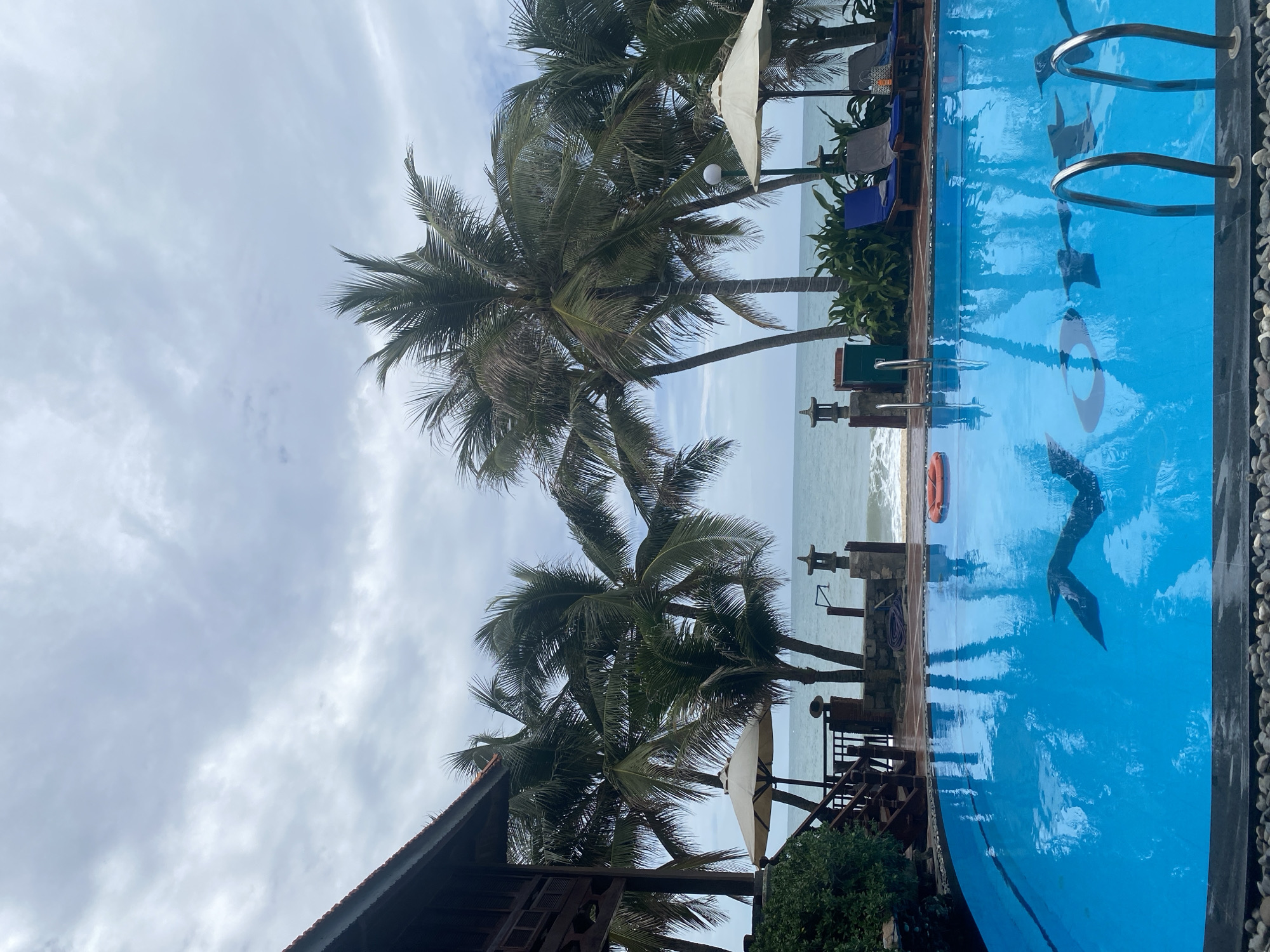 This media file captures a serene coastal resort scene in Phu Thuy Ward, Vietnam, featuring a vibrant blue swimming pool in the foreground. The image is taken on an overcast day, with a sky full of soft grey and white clouds, creating a diffused light over the landscape.
The swimming pool occupies the right side of the frame, its clear blue water reflecting the overhead sky and surrounding palm trees. Metal ladders provide access into the pool, and an orange lifebuoy floats near the edge. A subtle, circular design, possibly a letter 'O' or a logo, is visible on the pool's bottom, along with another abstract dark shape, resembling a stylized fish or wave pattern. The pool is bordered by dark coping stones and a light-colored pebbled area.
Beyond the pool, a lush line of tall palm trees with dense green fronds stands between the resort area and the ocean. Amongst the palm trees, several beige sun umbrellas and lounge chairs, some draped with blue towels, are arranged, suggesting an inviting space for relaxation. Stone lanterns or decorative elements are strategically placed along the pool's edge. To the far left, part of a wooden building with a tiled roof is visible, blending with the natural surroundings.
In the background, the calm ocean stretches to the horizon, its surface a soft blue-grey under the cloudy sky, with gentle waves breaking near the shore. No people are visible, emphasizing the tranquility and pristine nature of the resort area. The scene evokes a peaceful and relaxing holiday atmosphere.