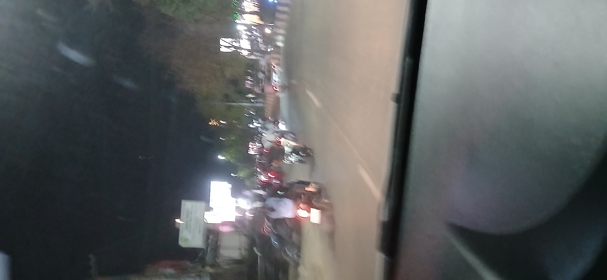 The image shows a street scene at night, likely in Samarinda, Indonesia, as indicated by the context. The foreground is dominated by the interior of a vehicle, possibly a dashboard, with a dark, textured surface. The background is blurry due to motion or low light conditions, typical of a night-time street view from a moving car.
Several motorcycles are visible on the road, some with riders. The headlights and taillights of the vehicles create streaks of light, indicating movement. A person wearing a light-colored shirt is clearly visible on a motorcycle in the mid-left of the image. There are also signs and buildings visible on the side of the road, some illuminated by streetlights or vehicle lights. Trees and other foliage are present, suggesting an urban or semi-urban environment. The overall impression is a busy street at night, with traffic and urban elements.