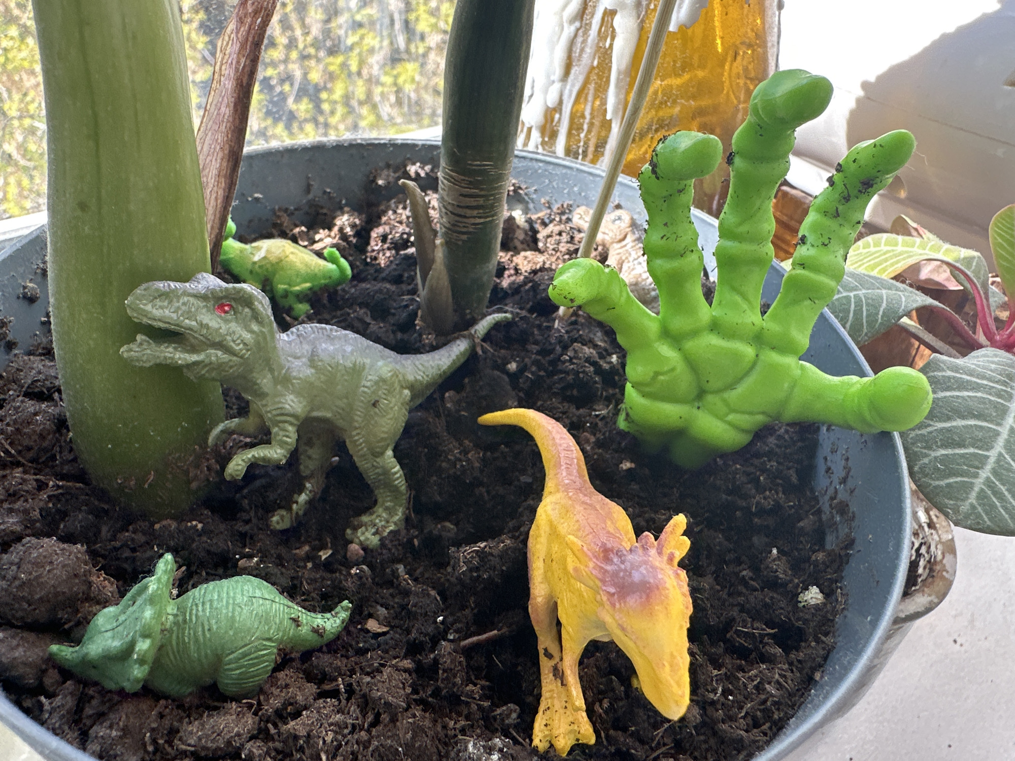 The image shows a grey pot filled with dark soil. Several objects are placed within the pot. There are at least five toy dinosaurs scattered in the soil: a grey tyrannosaurus rex, a yellow/orange dilophosaurus, two green triceratops, and one additional green dinosaur. A green monster hand with four fingers is also in the pot. A green plant stem rises from the soil in the back left of the pot. Other plants are also planted in the pot. The location of this image in Khimki, Russia cannot be confirmed by the details in the image.