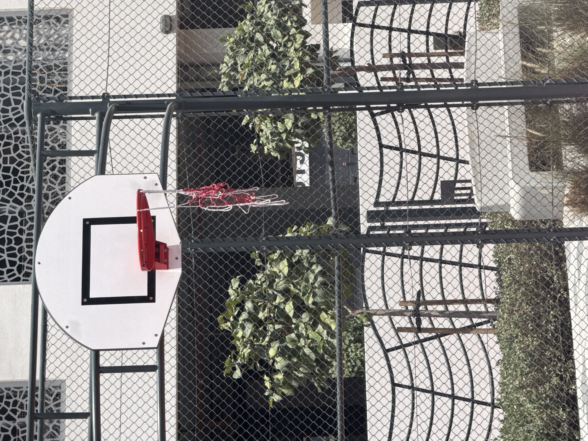 A basketball hoop is seen through a chain-link fence. The hoop is white with a black square outline, and a red rim. A red and white net is attached. Behind the fence, there is a white building with architectural details, including decorative panels. Some greenery is visible through the fence, appearing as small trees or shrubs. The location may be in Dubai, United Arab Emirates, but this cannot be confirmed solely from the visual content of the image.