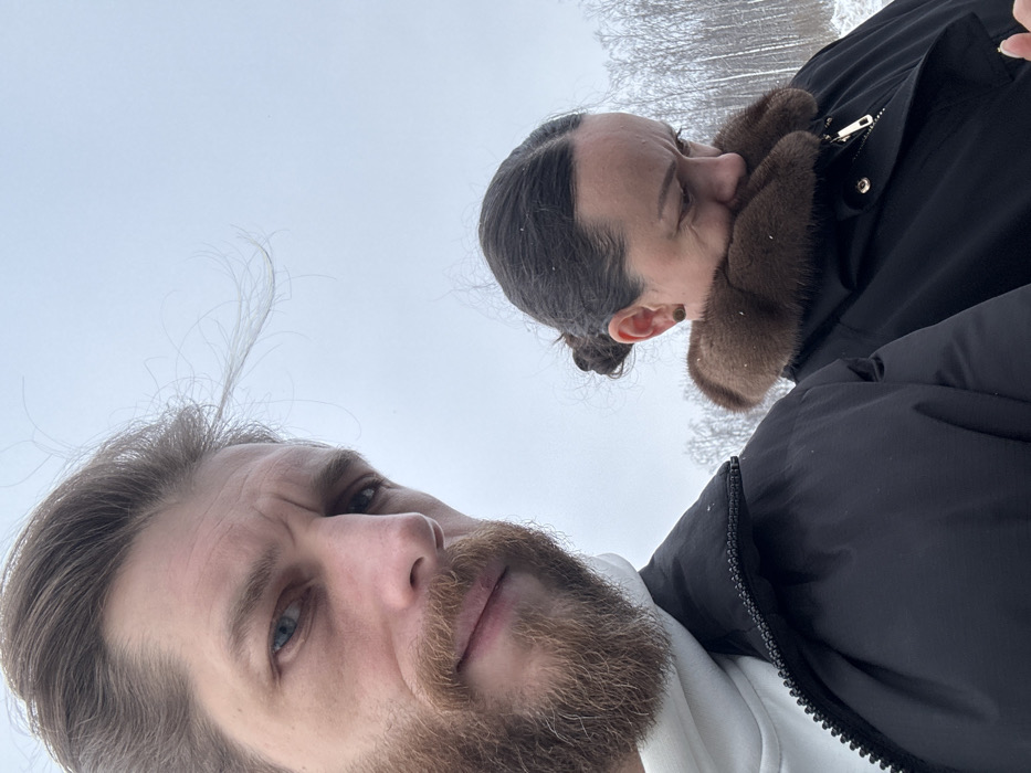 The image features two individuals, an adult male and an adult female, outdoors in a winter environment within the city of Novogorelovo, Russia. The male, positioned in the foreground on the left, has light skin, blue eyes, a light brown beard, and light brown hair showing signs of being windswept. He wears a white hooded garment beneath a black puffer jacket and is looking directly at the camera with a slightly furrowed brow. The female, situated to the right and slightly behind the male, has dark hair styled in a bun and wears a black winter coat with a prominent brown fur collar, partially obscuring her face. She is looking downwards and to her right, away from the camera, with a pensive expression. A small, dark stud earring is visible in her left ear. The background consists of a stark, light grey sky typical of an overcast winter day, with faint white specks indicating light snowfall. A dense line of bare deciduous trees, primarily white-trunked, is visible along the horizon, suggesting a natural or wooded area.