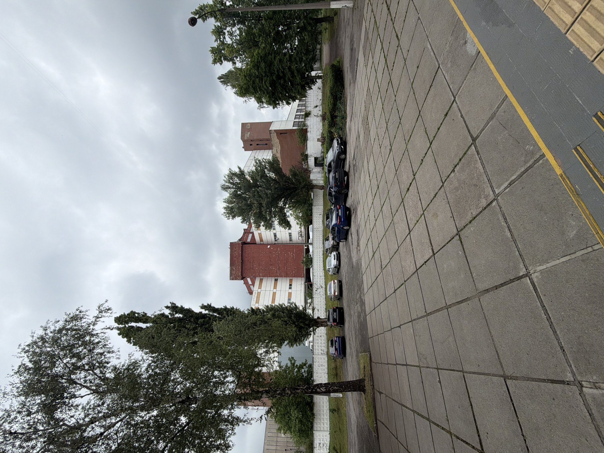 The image shows a courtyard with a large tiled area in the foreground. Several cars are parked in the background, near a long, white, tiled wall. Behind the wall, there is a multi-story building with red and white sections. Tall trees are scattered around the courtyard, and a street lamp is visible on the right side of the image. The sky is overcast and gray. This appears to be a commercial or industrial area.