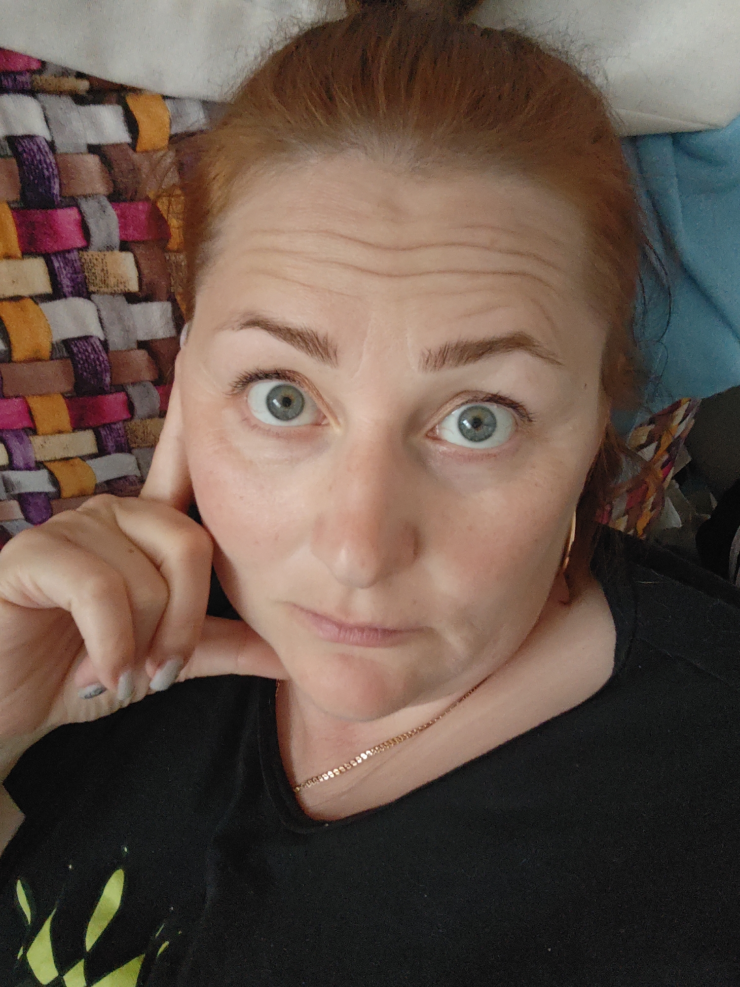 A fair-skinned woman with auburn hair pulled back, light blue eyes, and freckles is shown in a close-up, selfie-style photograph. Her right hand is raised to her face, with fingers touching her cheek. She is wearing a black t-shirt and a gold necklace. The background shows a multi-colored woven textile on one side and a blue surface on the other. The location is unknown, and there is no evidence connecting the image to Markovskii, Russia.