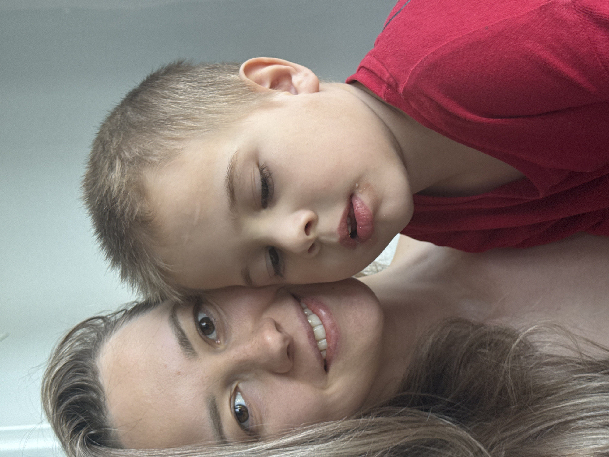 A young boy in a red shirt rests his head on a woman with long, light brown hair. The woman is smiling. The location is unknown. There is no information to indicate that the image was taken in Chaikovsky, Russia.
