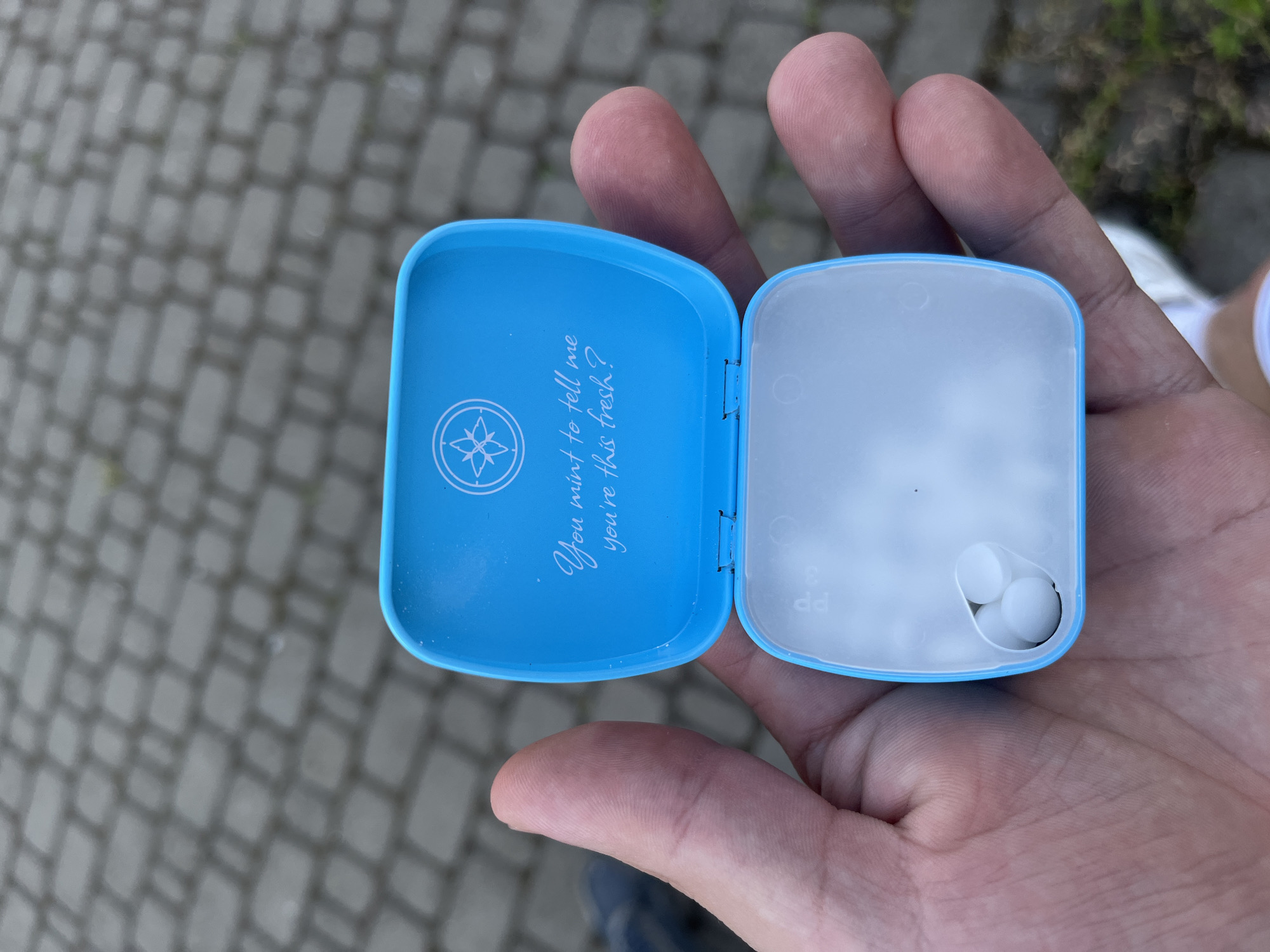 A person's hand holds an open, light blue tin of mints. The tin is square with rounded corners. The lid, on the left, features a white compass-like logo and the handwritten text "You mint to tell me you're this fresh?". The interior of the lid and the bottom of the tin are white. The bottom of the tin contains a few small, white, round mints. The background is a gray paved surface, possibly a sidewalk, with some green grass visible on the right. The lighting suggests it is daytime. The image was taken in Kyiv, Ukraine.