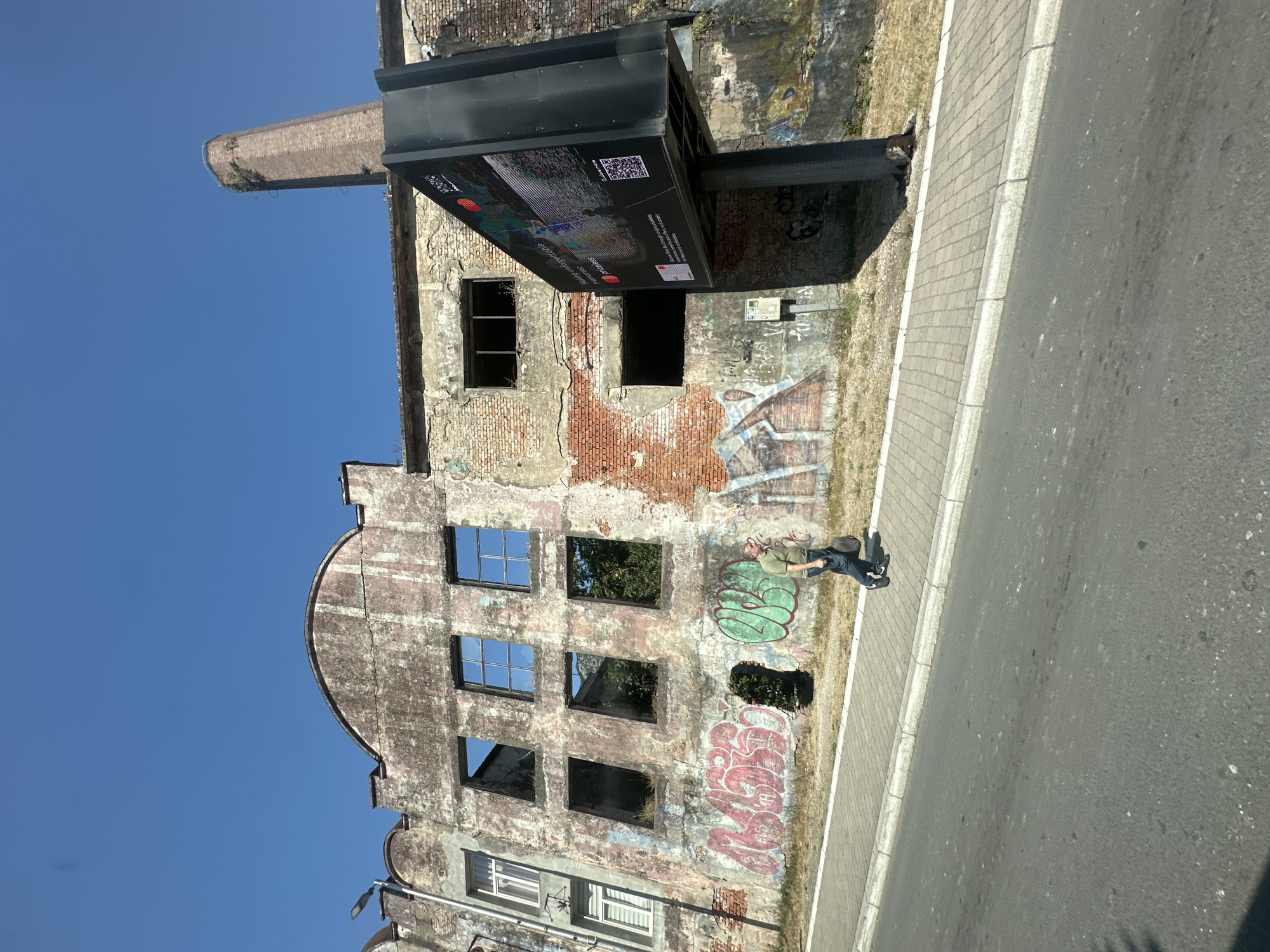 A man walks along a sidewalk next to a derelict building in Škaljari, Montenegro. The building, made of brick and plaster, is in disrepair with broken windows and graffiti. A large billboard stands in front of the building. The sky is clear and blue.