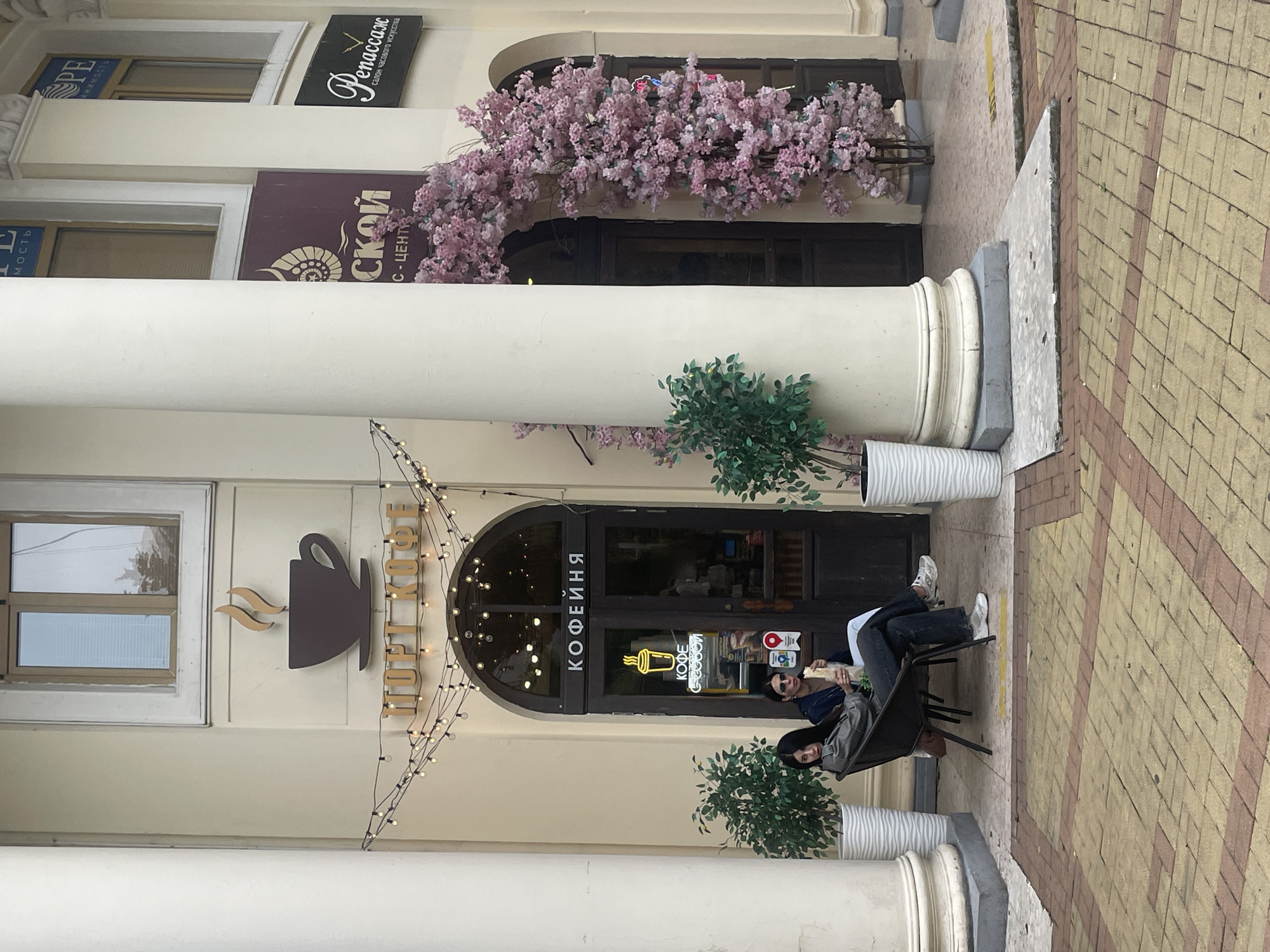 The image shows a coffee shop in Sochi, Russia. The building has a light beige facade with white columns. Over the entrance, there is an arched doorway, a sign with a coffee cup, and string lights. Above the coffee shop are signs in Russian with decorative elements. Pink floral decorations adorn the upper section of the building. Two women are seated in chairs outside the coffee shop. The street is paved with light brown bricks. Potted green plants are placed near the entrance of the coffee shop.
