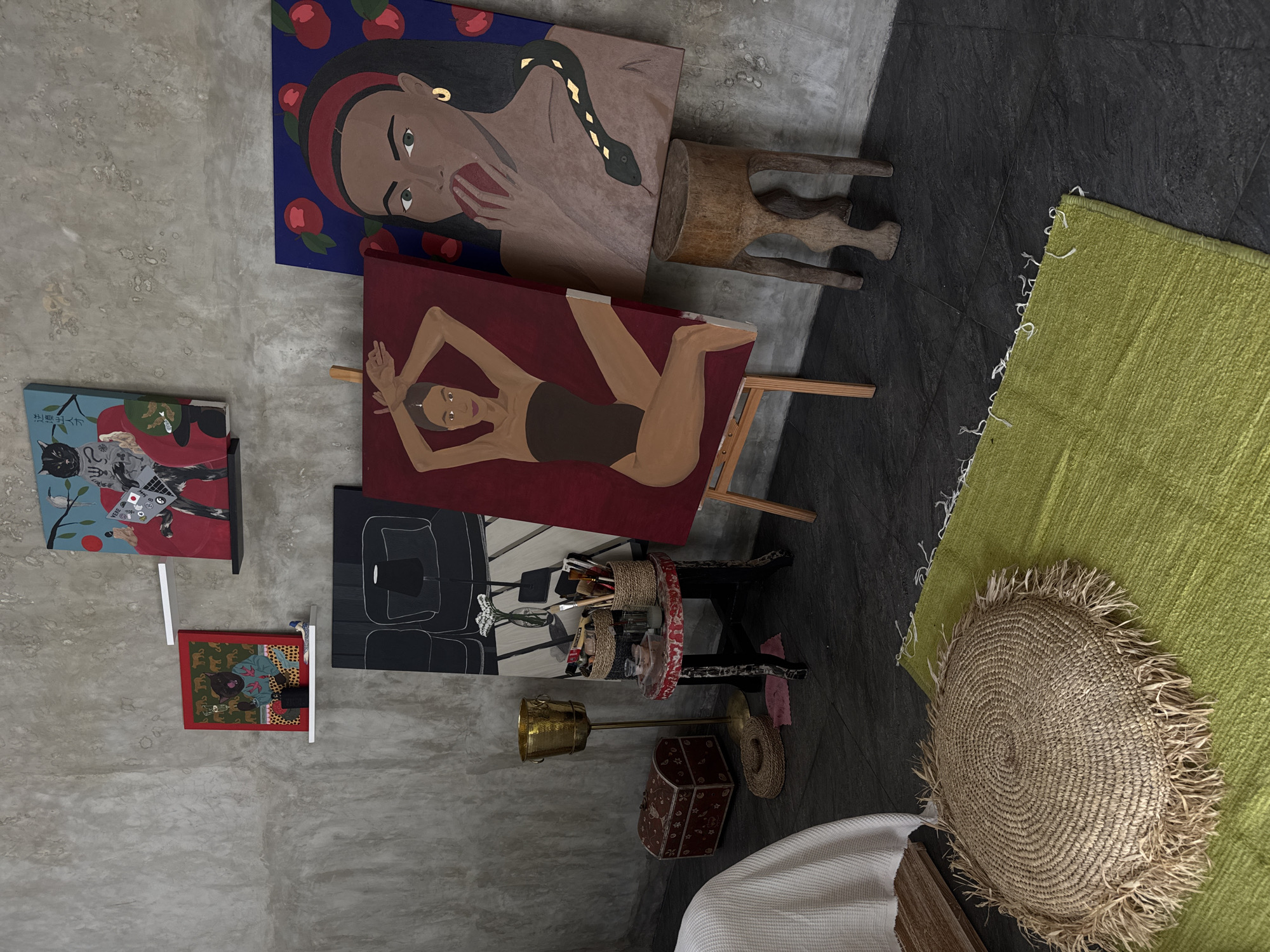 The image depicts an interior space in North Kuta, Indonesia. The room has a modern, artistic aesthetic. Several paintings are displayed on the wall, including portraits of a woman with apples and a snake, and another of a woman in a swimsuit. Other artworks present abstract and cartoonish designs. A wooden stool and an easel are present. On the floor, there is a green rug, a round straw mat, a sewing table, a brass standing ashtray, and a decorated wooden box, all arranged in a semi-organized manner. The floor is made of a combination of grey and black stone tiles. Part of a white bed or seating area is visible in the lower right corner of the frame. The walls have a rough, concrete texture.