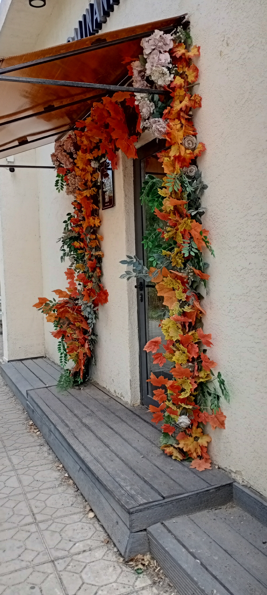 The image shows an exterior view of a building entrance decorated with artificial autumn foliage. The foliage, primarily in shades of orange, yellow, and red leaves, is draped around the doorway and under an awning. Some artificial flowers, in muted tones of pink and grey, are also incorporated into the decoration. The entrance features a glass door and a textured cream-colored wall. To the left of the entrance, there is a wooden ramp or deck made of gray planks. The ground in front of the ramp is paved with interlocking concrete tiles. The overall scene suggests a decorative arrangement for the fall season, likely outside a commercial establishment. There are no people visible in the image.