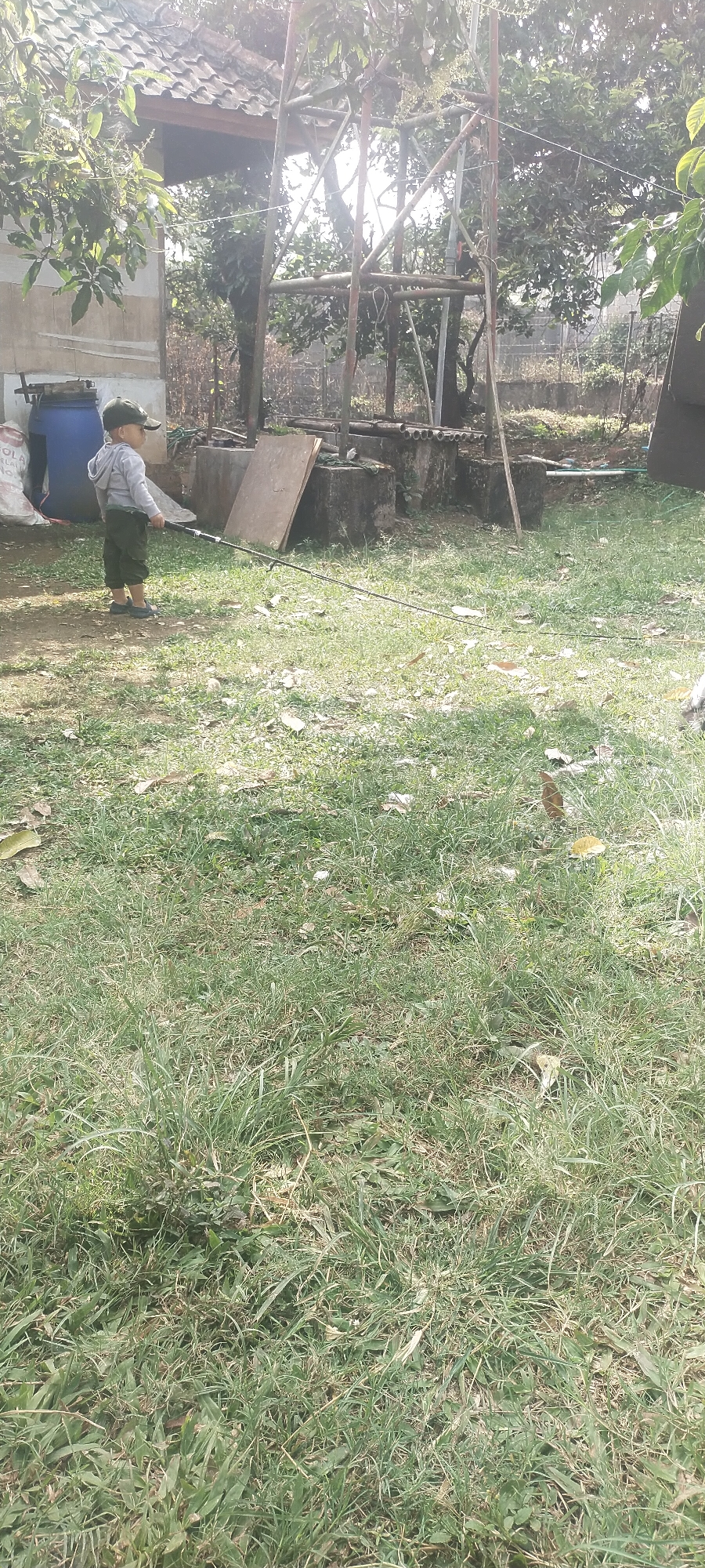 In Pasirhuni, Indonesia, a young child, dressed in a gray hoodie and green pants, stands in a grassy outdoor area, holding a fishing rod. The child is facing away from the camera and appears to be engaged in fishing. In the background, a blue barrel and a large blue bag are visible. A metal structure, possibly a water tower, is also present, along with trees and a tiled roof of a building. The ground is covered in grass, with scattered leaves and shadows from overhead sunlight indicating it is daytime. The overall environment suggests a rural or semi-rural setting within the city.