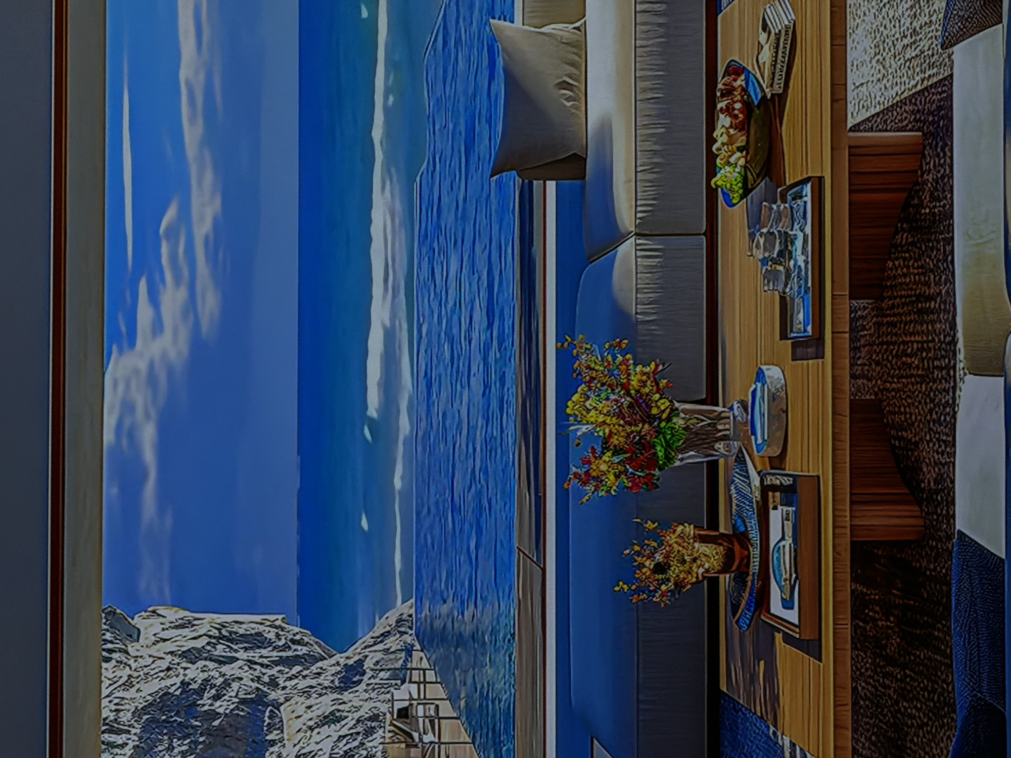 The image displays a scene that appears to be a landscape photograph rotated 90 degrees clockwise. When viewed in its intended landscape orientation (rotated 90 degrees counter-clockwise), it shows a luxurious indoor living space with a panoramic view of an outdoor scene.
**Subjects:**
The foreground (right side in corrected orientation) features a comfortable, light-colored sofa with a matching cushion. In front of the sofa is a wooden coffee table, adorned with several items: bowls filled with snacks such as grapes and nuts, a tray with glasses, two vases holding colorful flower arrangements, and a framed picture or decorative object. No people are visible.
The background (left side in corrected orientation) presents a dramatic outdoor vista, likely a large window, a digital screen, or a mural. It depicts a clear blue sky with fluffy white clouds, a vast blue ocean or sea with visible waves, and in the lower left corner, rugged, snow-capped mountains or rocky cliffs.
**Activity or event taking place:**
There is no direct activity depicted as no individuals are present. The arrangement of snacks and drinks on the coffee table suggests the setting is prepared for relaxation, entertaining, or leisure.
**Notable details:**
The scene conveys a bright, daytime atmosphere, indicated by the clear sky and what appears to be natural light. The indoor setting is modern and well-appointed, implying comfort and luxury. Given the provided location context of Sofiivska Borshchahivka, Ukraine, which is landlocked, the expansive ocean and mountain view is almost certainly not a real window view but rather a very large painting, a high-resolution display, or part of the interior design itself. The overall mood is tranquil and inviting.
**Visible text:**
There appears to be some unidentifiable text on one of the items on the coffee table, possibly a book or decorative box, but it is not legible.