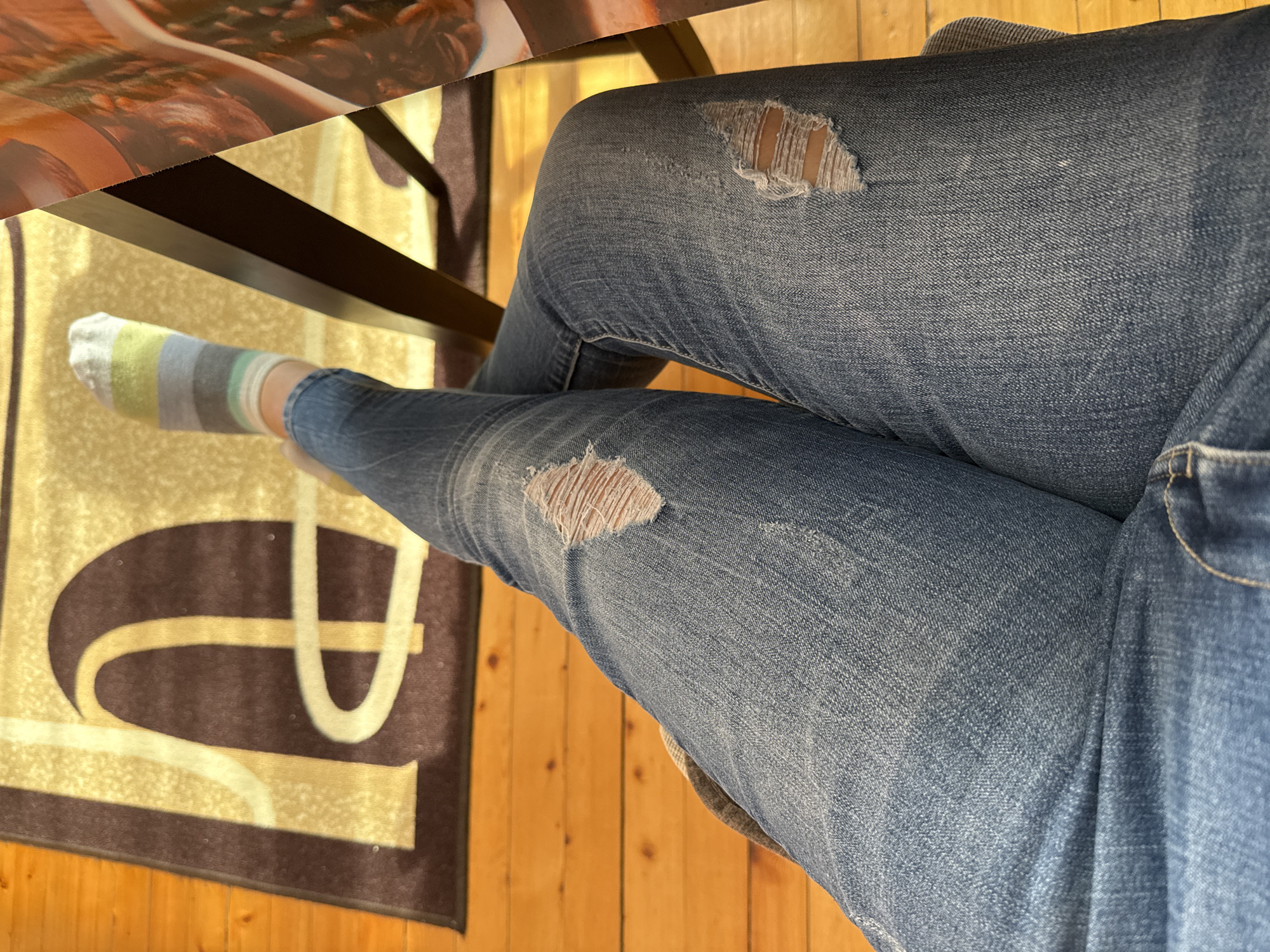 The image depicts a person sitting at a table. The person is wearing ripped blue jeans and a striped sock. The table has a dark brown leg and a surface with a pattern that is only partially visible. Beneath the table, there is a rug with a brown and tan design. The floor is made of wooden planks. There are no indications to determine if the image was captured in Sofia, Bulgaria.