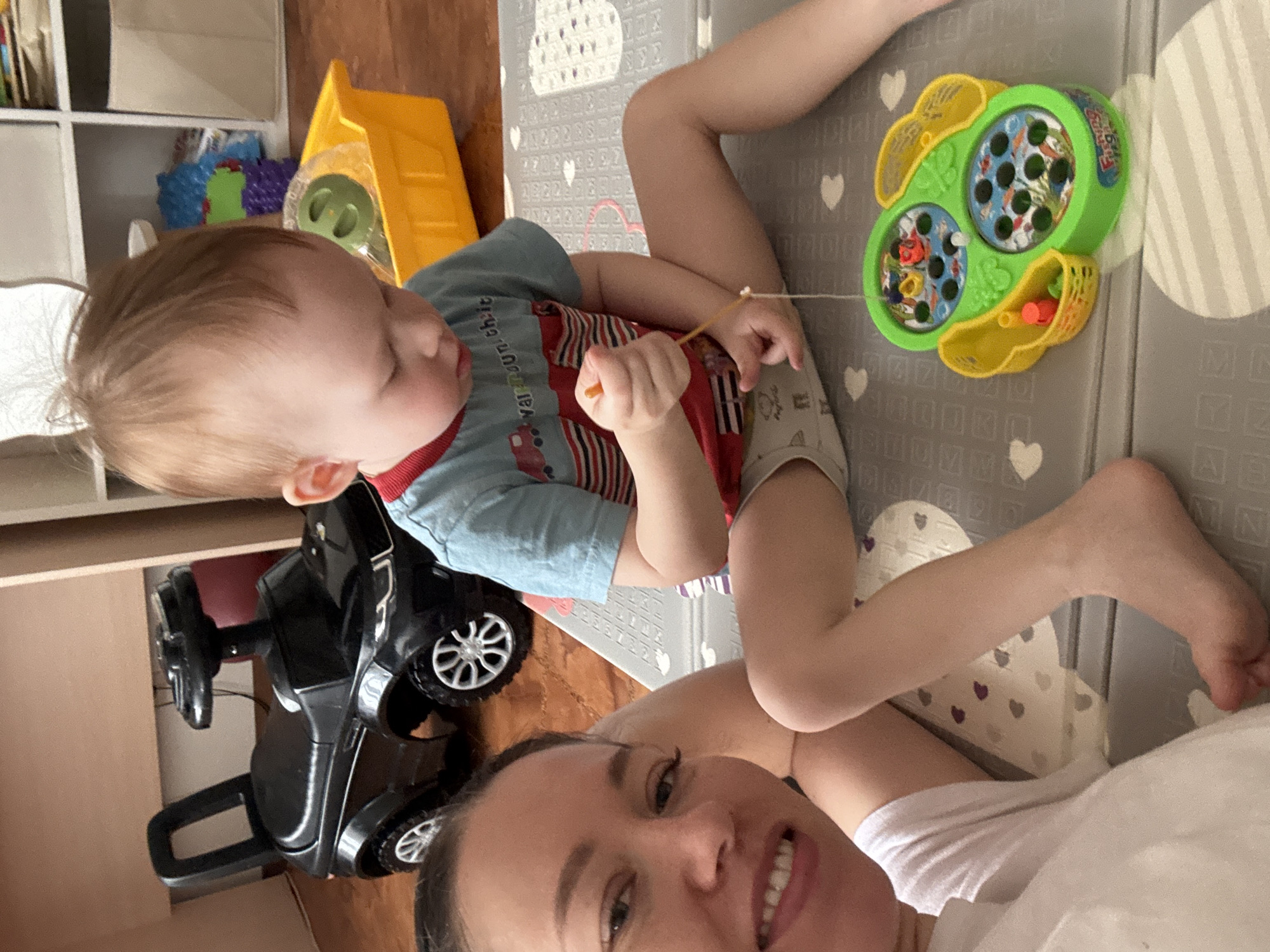 A selfie taken indoors shows a woman lying on her side, smiling at the camera. Beside her, a young child, about one year old, is seated on a gray playmat with white hearts and geometric shapes. The child is playing with a toy fishing game; the toy is green and yellow with small fish inside. A yellow plastic toy truck is in the background, along with a shelf. A black toy car is also visible near the woman and child. The setting appears to be a home playroom or living area. There is no information available in the image to confirm the location in the city of Ferma, Russia.