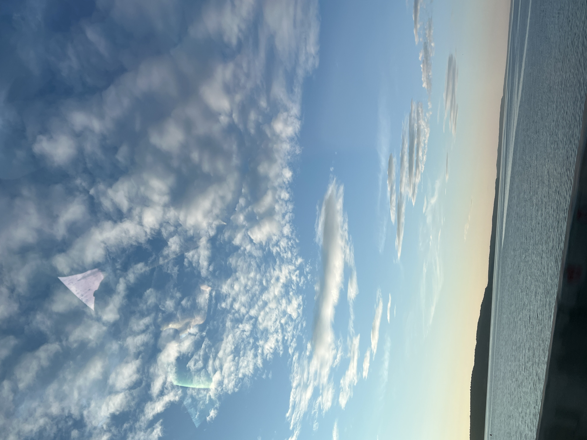 The image depicts an expansive landscape captured from an elevated perspective, likely from a vehicle traversing a bridge, overlooking a substantial body of water under a partially cloudy sky. The location is the city of Chaikovsky, Russia, specifically consistent with a view over the Votkinsk Reservoir or Kama River.
The upper two-thirds of the image are dominated by the sky, displaying a clear gradient from a pale blue-yellow near the horizon to a deeper azure overhead. Various cloud formations are present: dense, white cumulus clouds are scattered across the upper sky, while the mid-sky features more dispersed, smaller, and some elongated cloud wisps. Prominent reflections of light and objects (including a distinct light purple, triangular shape and colorful lens flare-like streaks) are visible in the upper-left quadrant of the sky, indicating the photograph was taken through a window.
The lower third of the image features a calm, blue-green body of water, showing subtle ripples across its surface. A low, dark, forested landmass delineates the distant horizon, suggesting a shoreline or island across the water. A dark grey concrete barrier or railing runs horizontally across the bottom of the frame, separating the foreground from the water, consistent with a bridge structure. A small, dark, possibly buoy-like object is faintly visible on the water's surface near the distant left shore. The soft, warm light near the horizon suggests either early morning or late afternoon. No people, specific vehicles (other than the one from which the photo was taken), or distinct man-made structures are visible on the water or distant landmass.