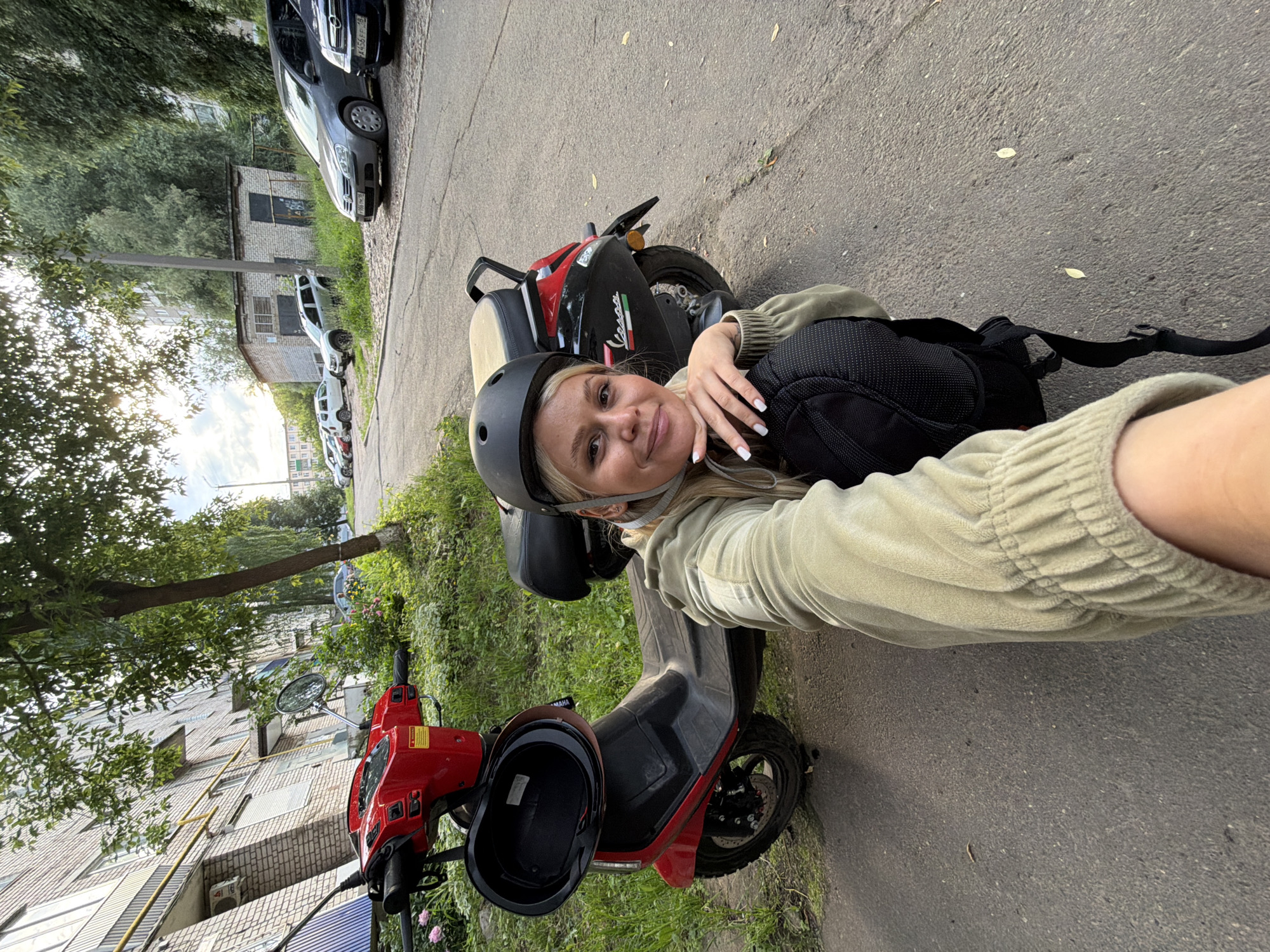 A woman wearing a black helmet and a light olive green hooded jacket kneels beside a red and black scooter on an asphalt road. The scooter has a black helmet resting on its front. A black backpack is on the ground next to the woman. In the background, there is a multi-story apartment building with a yellow pipe running along its facade. Trees line the street, and several cars are parked along the side of the road. The setting appears to be a residential area in the city of Sarapul, Russia, likely a courtyard or street.