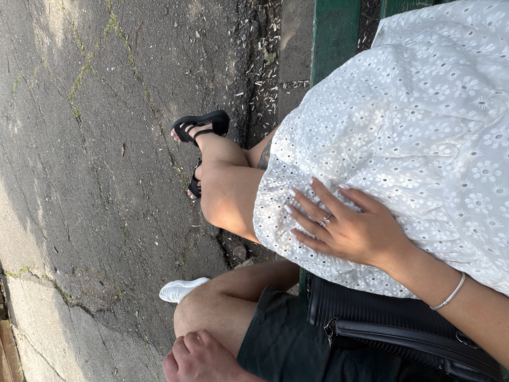 An elevated view in Sofia, Bulgaria, depicts two individuals seated on a green bench in an outdoor urban setting. The person on the left is partially visible, showing a right thigh in dark green shorts, a left forearm and hand resting on the right thigh, and a right foot wearing a white athletic sneaker. The person on the right wears a white eyelet (broderie anglaise) dress. Their left hand, adorned with a silver ring featuring two pink butterfly-shaped stones and manicured nails with a white iridescent finish, rests on the dress. A silver snake-chain bracelet is visible on their right forearm. Their left foot is visible, wearing a black platform sandal, and a faint tattoo is present on their inner left thigh. A black structured handbag with a shoulder strap is positioned on the bench next to this individual. The ground is a cracked asphalt or concrete surface with interspersed patches of grass and weeds, along with scattered debris including small organic matter and white cylindrical objects resembling cigarette butts. The scene is illuminated by direct sunlight, casting distinct shadows.