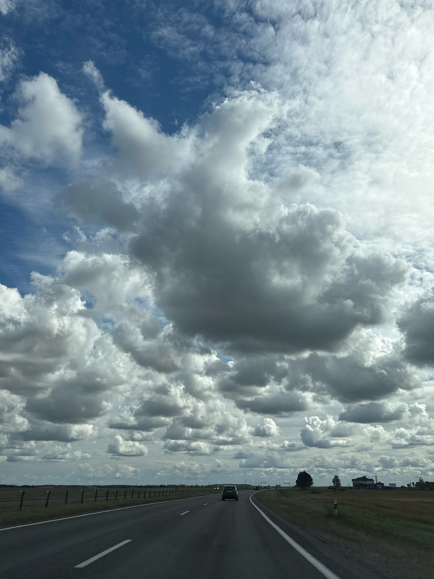 The image depicts a view from a vehicle traveling on an asphalt road in Блонскі сельскі Савет, Belarus. The road is straight and curves slightly to the right in the foreground, with white lane markings. To the left of the road is a flat, grassy area bordered by a fence. To the right of the road, there is more flat, grassy terrain with some scattered trees and buildings in the distance. The sky is filled with a dramatic display of clouds. Large, cumulus clouds with dark undersides are interspersed with brighter, wispy cirrus clouds against a blue sky. The lighting suggests it is daytime, with the sun intermittently breaking through the cloud cover, casting shadows on the landscape. A dark-colored car is visible on the road ahead, traveling in the same direction as the viewer. The overall scene conveys a sense of a journey through a rural Belarusian landscape under a dynamic sky.