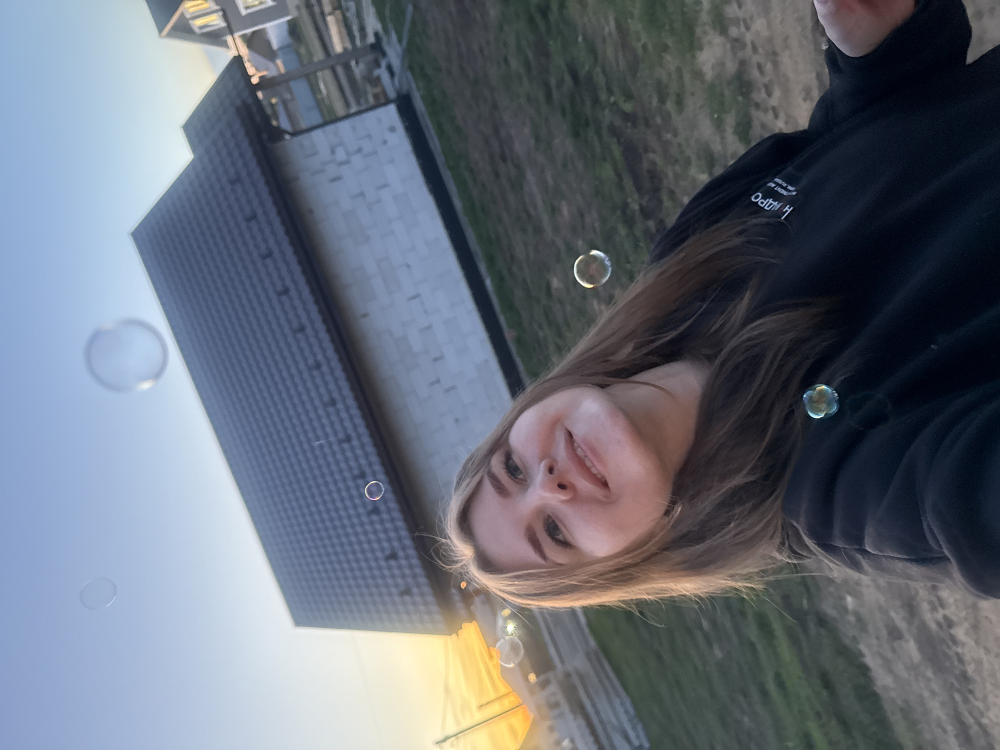 A young woman stands outside in the city of Ферма, Russia. She is wearing a black hooded sweatshirt. Several bubbles are floating in the air around her. In the background, there is a building with a dark roof and light-colored siding, and a grassy area. The sky is a pale blue-orange color.