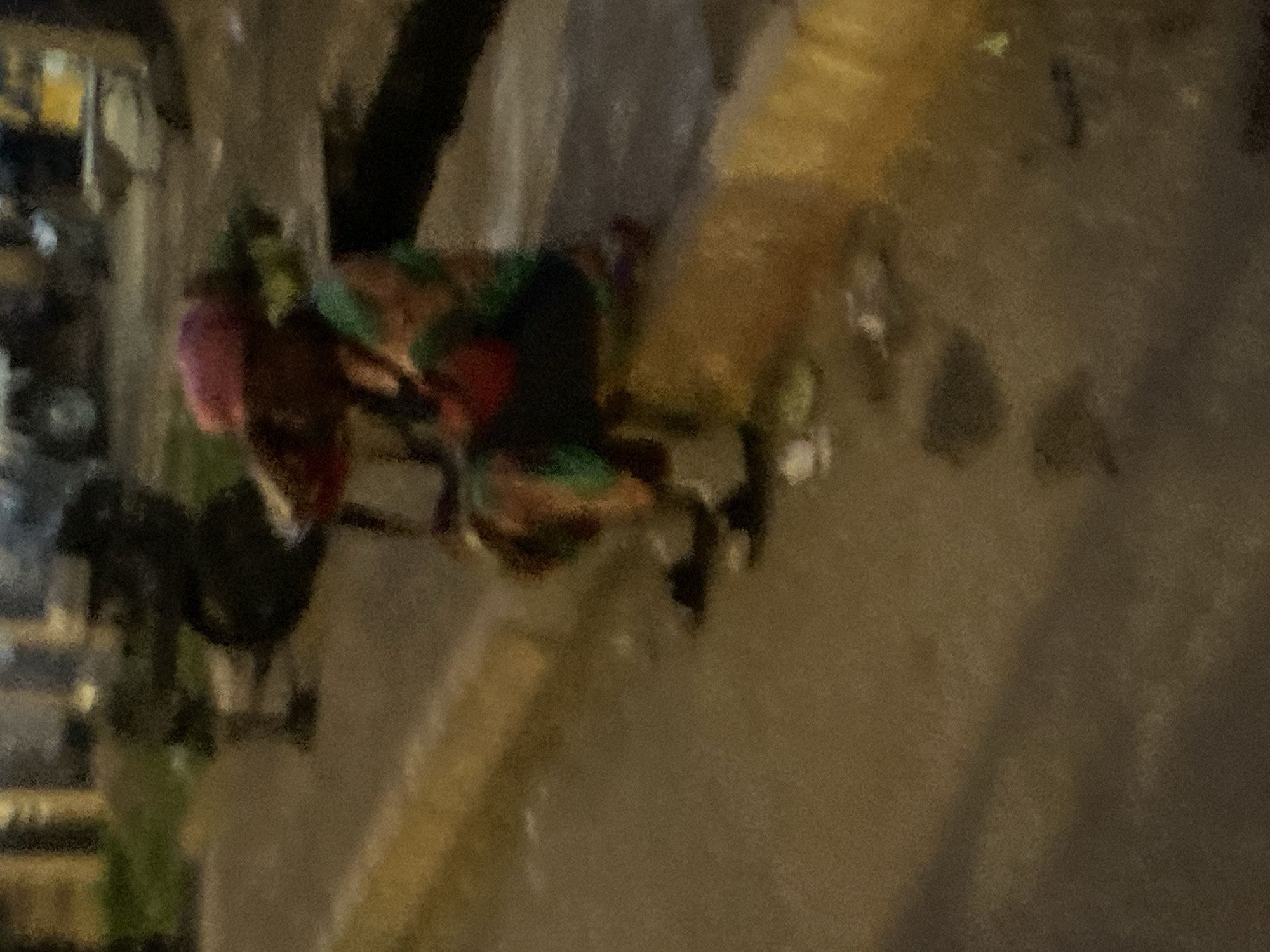 This blurry, low-light image depicts an outdoor street scene in Bukuru, Nigeria, likely during the evening or night.
In the mid-ground right, a person is seated on a raised, yellowish-brown concrete curb. This individual wears a pink head covering, a multi-colored garment featuring green, red, and dark patterns, and dark footwear, possibly sandals. A small red bag or object is visible on their lap or chest area. Several small, light-colored pieces of debris or litter are scattered on the dark, paved ground directly in front of and around the seated person's feet.
Further into the blurry background on the left, two indistinct figures are visible, appearing to stand or walk. The background contains extremely blurred shapes suggestive of buildings and vehicles, with several diffused points of light indicating artificial illumination. A faint patch of green vegetation can be discerned behind the figures on the left. The overall environment suggests a common urban or semi-urban setting with pedestrian and vehicular presence.
