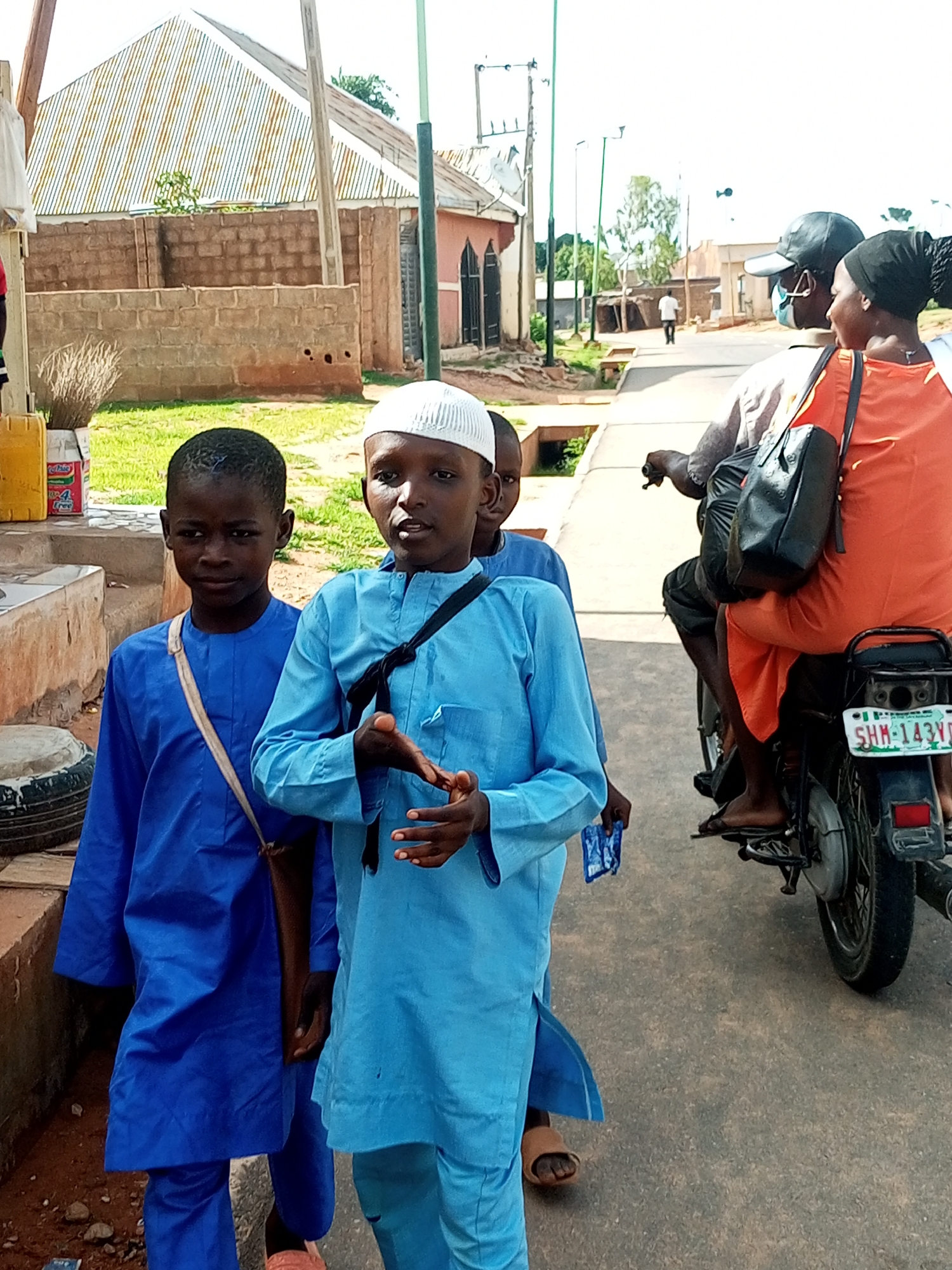 The image depicts a street scene in Tempure, Nigeria. In the foreground, two young boys are standing. The boy on the left is wearing a bright blue kaftan and a dark cross-body bag. He has short dark hair and is looking to his left. The boy on the right is wearing a lighter blue kaftan and a white skullcap. He is gesturing with his hands as if speaking and looking forward. Behind them, a person is riding on the back of a motorcycle, wearing an orange garment and a black headscarf. The driver of the motorcycle is wearing a black cap and a face mask. The motorcycle has a license plate that reads "SHM-143YD". The background shows buildings with corrugated metal roofs and brick walls, as well as streetlights and some vegetation. The ground is paved.