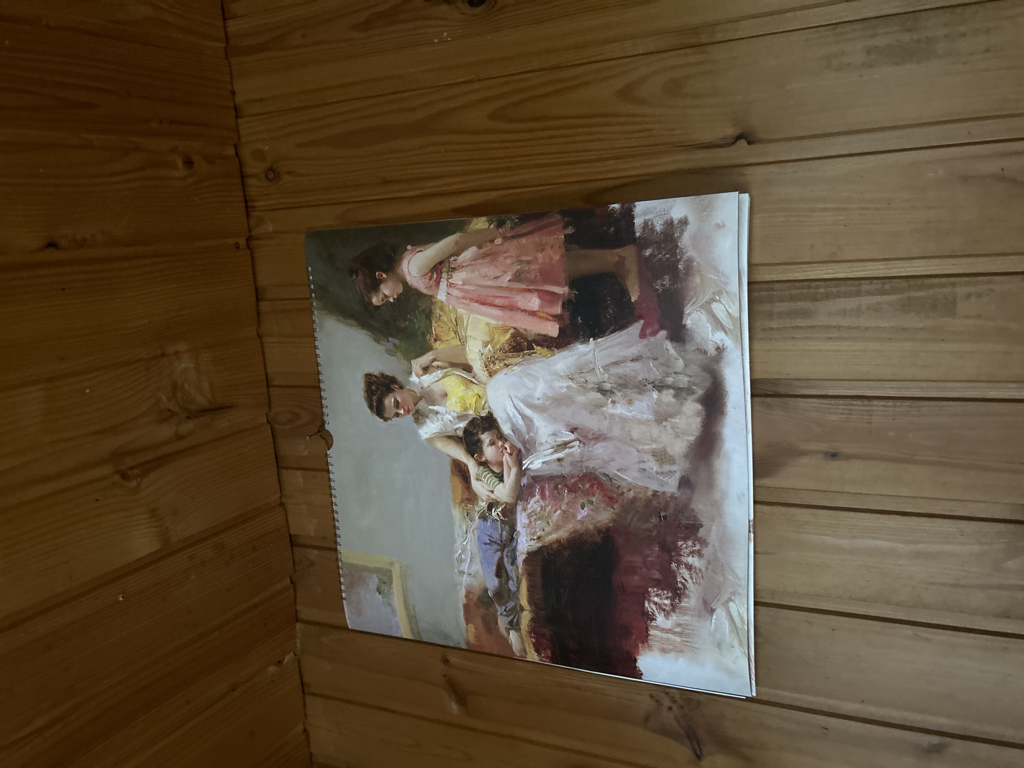 A painting hangs on a wooden paneled wall. The painting depicts a mother with two children. The mother is seated, wearing a white dress and a yellow bodice, and is holding a sleeping child on her lap. The child is wearing a blue jumpsuit and a striped shirt. To the right of the mother, a young girl stands wearing a pink dress. The background of the painting is a dimly lit interior scene, possibly a room. The wooden paneled wall suggests an indoor setting, possibly a home or cabin. The image does not contain any information about the city of Leningradskaya, Russia.