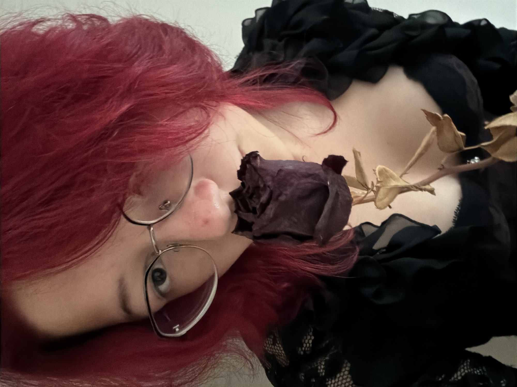 The image shows a close-up, head-and-shoulders view of a person with bright red hair and glasses. The person is holding a dried, dark purple rose near their face. They are wearing a black garment with lace detailing and ruffled accents around the neckline. The background is a plain, light-colored wall. The scene is indoors, with soft lighting. There is no indication of the location being in Chelyabinsk, Russia or any other specific city.