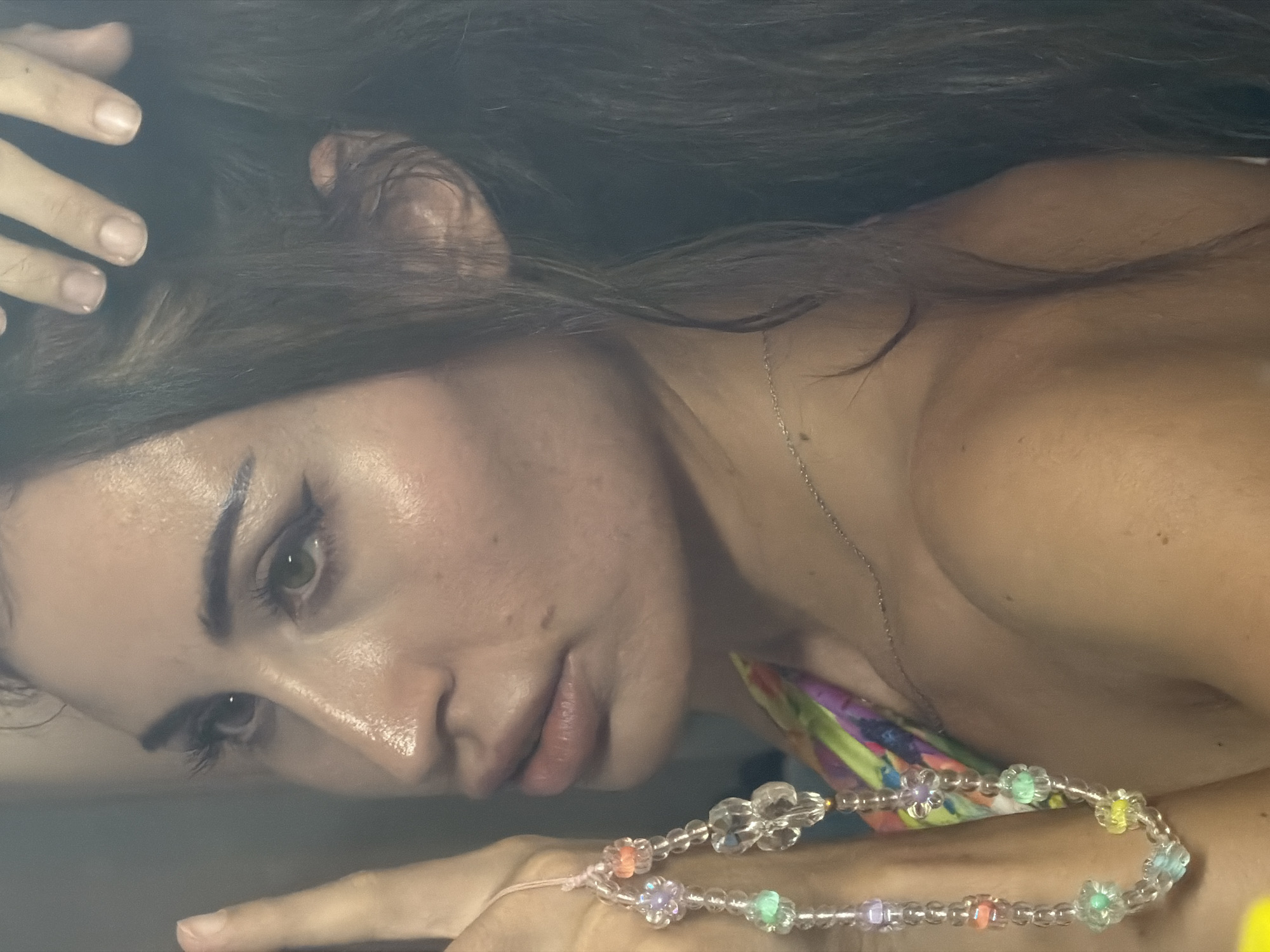 A young woman, likely in her early to mid-twenties, is captured in a close-up, waist-up portrait. She is looking to her left, with her right hand touching her hair. She is wearing a colorful, patterned halter top and a delicate silver necklace. A beaded phone charm hangs from her left hand. The background is indistinct and appears to be indoors, possibly a bathroom or bedroom, with a mirror reflecting the scene. The lighting is soft and diffused, creating a slightly hazy effect. The image does not provide any specific details about the location in Tersefanou, Cyprus.