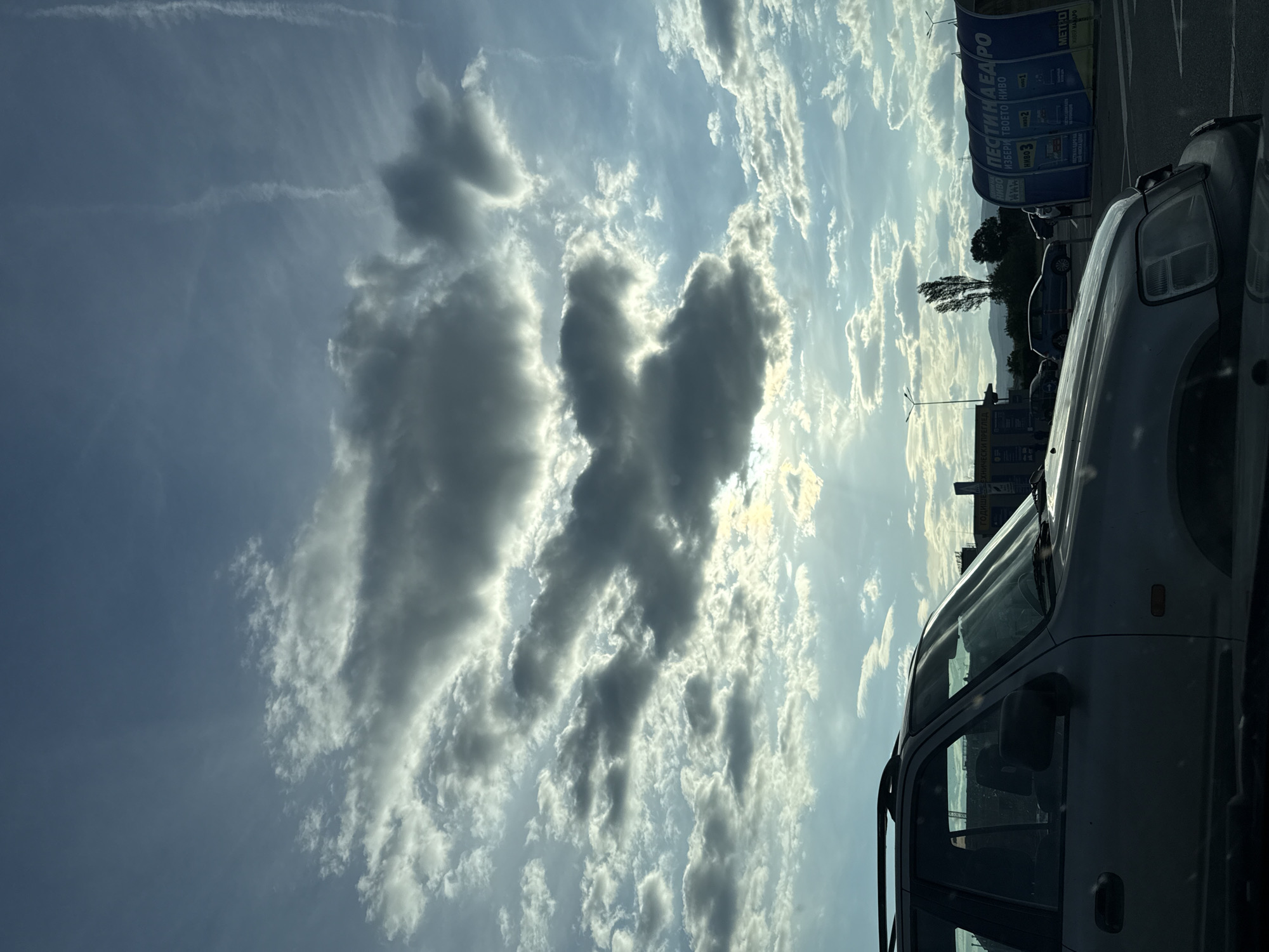 The image shows a view of the sky with scattered clouds, including a large cloud formation in the center resembling a stylized butterfly or two joined hearts. A light aircraft contrail is visible in the upper left.
In the foreground, a silver or light-colored SUV is parked, occupying the right side of the frame. To the right of the car, an advertisement with the name "ПЕСТИНА НИВАРО" and "METPO" is visible.
In the background, the tops of buildings and a few trees are visible, along with utility poles. Based on the advertisement, the location is in or near Stara Zagora, Bulgaria, likely close to a METRO store.