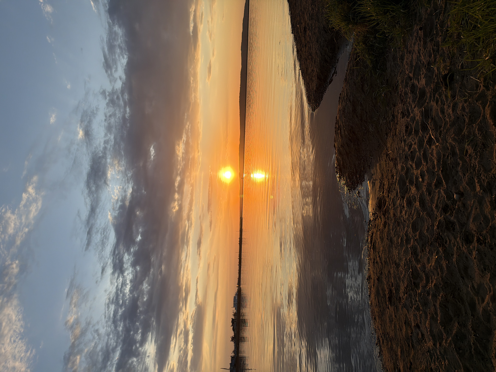 The image captures a sunset over a body of water, likely a lake or wide river, with the sun's reflection creating a vertical line of light in the water. The shoreline is visible on the right side, with sandy or muddy ground meeting the water's edge, and some vegetation growing near the bank. The sky is filled with clouds, ranging from light blues and whites to darker grays, reflecting the colors of the setting sun. The location is Unknown, Unknown.