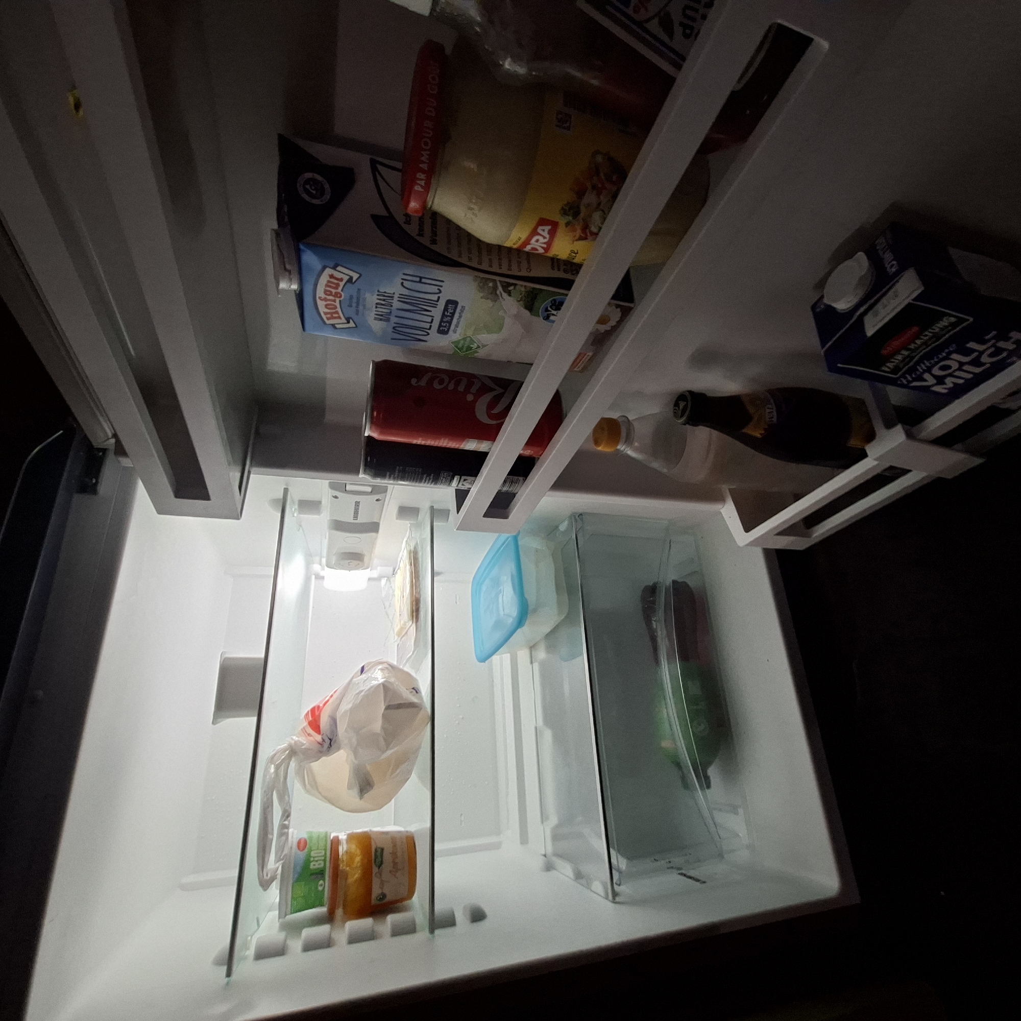 This media file shows the inside of a refrigerator in Rastatt, Germany, likely during daylight hours given the interior light. The refrigerator is moderately stocked with various food and drink items.
Prominent objects include several cartons of milk, specifically "Hofgut VOLLMILCH" and another dark blue carton labeled "VOLLMILCH" with "FAIRE HALTUNG" visible. There is also a red can of "River" soda and a dark glass bottle, possibly wine or beer, with a yellow cap. A glass jar with a red lid, labeled "PAR AMOUR DU GOUT," contains a light-colored spread. Other items include a blue plastic container, a clear plastic container holding a green bottle, and a plastic bag with a white handle containing food items. Smaller packages, one labeled "Bio," are also present on a lower shelf. The lighting within the refrigerator is illuminated by its internal light. The overall impression is of a typical home refrigerator interior, organized with various groceries.