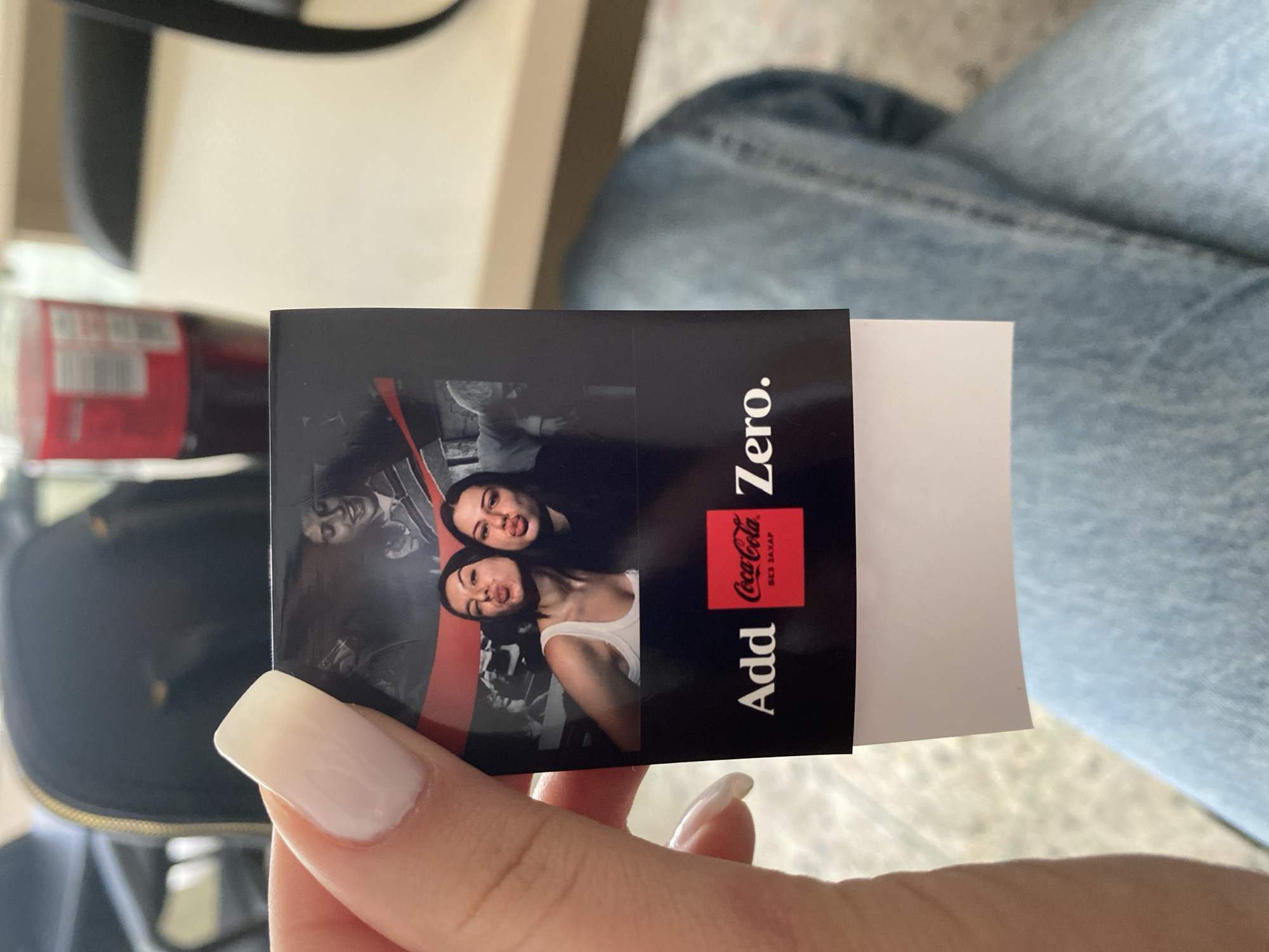 A person's hand, with manicured nails, holds a Coca-Cola Zero branded print in an indoor setting. The print features a black background with the Coca-Cola Zero logo, and the words "Add Zero." printed on it. The print also includes a black and white photo of two people making faces. The print appears to be a promotional item, likely dispensed from a vending machine, as part of a marketing campaign. The environment includes a part of a purse, a red Coca-Cola bottle, jeans, and a light beige table. The location is Plovdiv, Bulgaria.