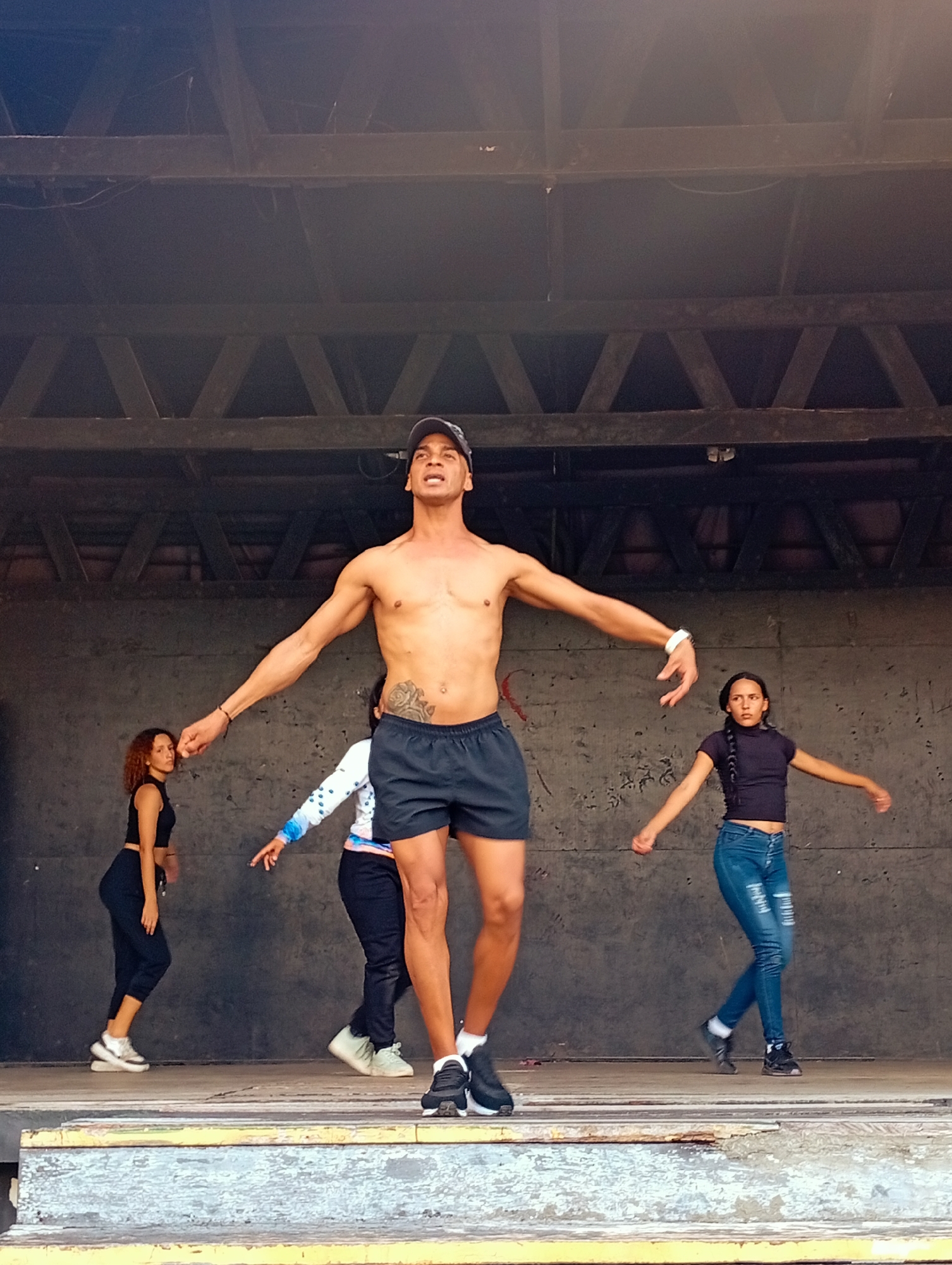 On a stage in Playa El Agua, Venezuela, a shirtless man in a baseball cap, black shorts, and black-and-white sneakers stands with his arms outstretched. He has a tattoo on his left abdomen. Three women are also on the stage, dancing. From left to right, the first woman wears a black crop top, black pants, and white sneakers. The second woman wears a white jacket with blue sleeves, black pants, and light-colored sneakers. The third woman wears a black long-sleeved shirt, blue jeans, and black sneakers. The stage has a weathered appearance, with multiple layers of paint visible. The background consists of a dark wall with a wooden support structure overhead.